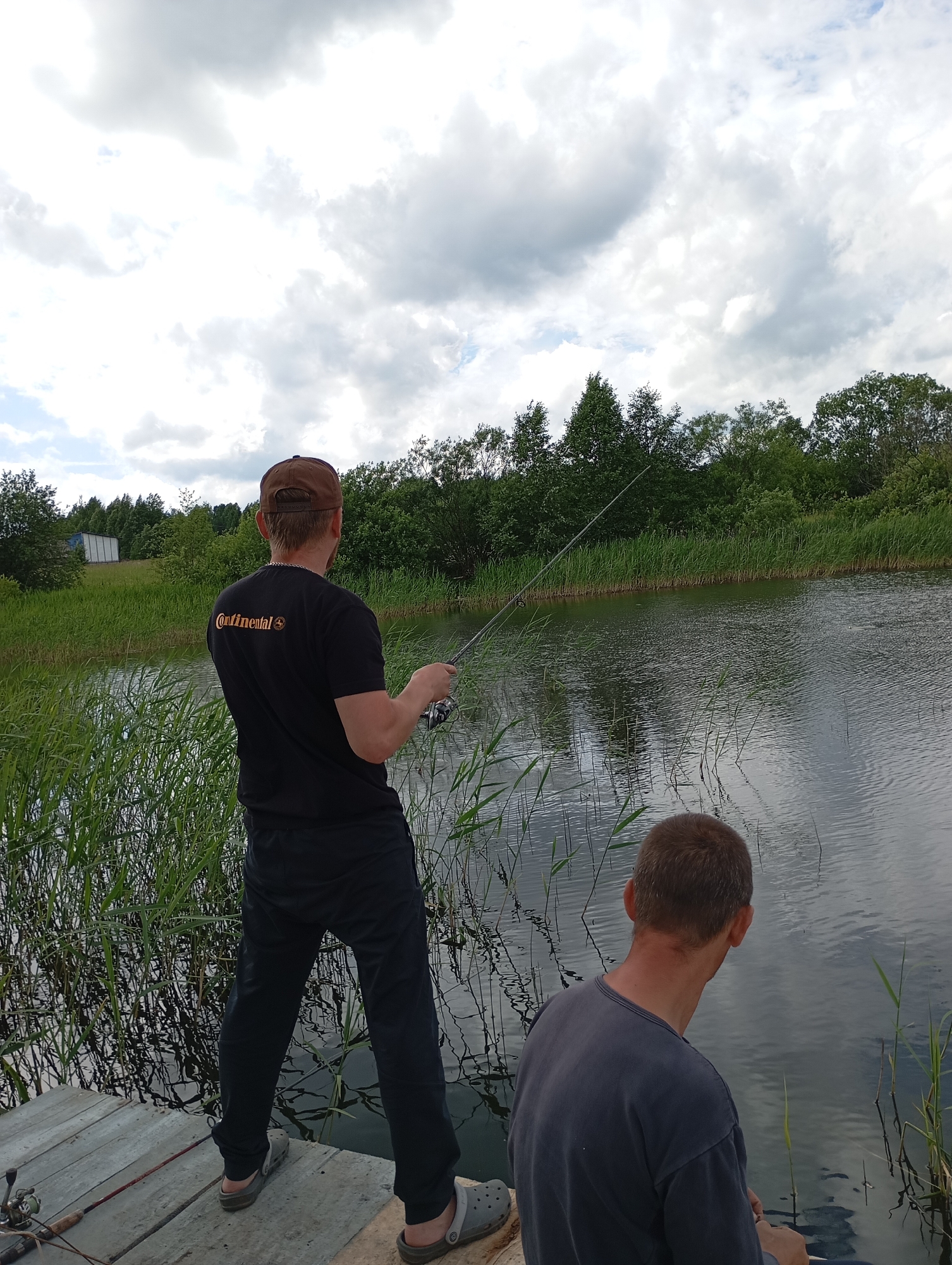 Two individuals are fishing on a wooden dock. The person in the foreground is seen from the back, wearing a dark blue long-sleeved shirt and dark pants, with their head turned to the right, facing the water. The second person, also seen from behind, is wearing a black t-shirt with the "Continental" logo and a brown baseball cap. They are actively fishing, holding a fishing rod and reel. The dock is made of weathered wooden planks. Tall green reeds and grass line the edge of the body of water. In the background, a dense line of green trees and bushes stretches across the horizon. A white building with a metallic roof is visible in the distance to the left, amidst the greenery. The sky is overcast with scattered clouds. The overall scene depicts a serene fishing activity in a natural, rural environment, likely the village of Ryabki, Russia.