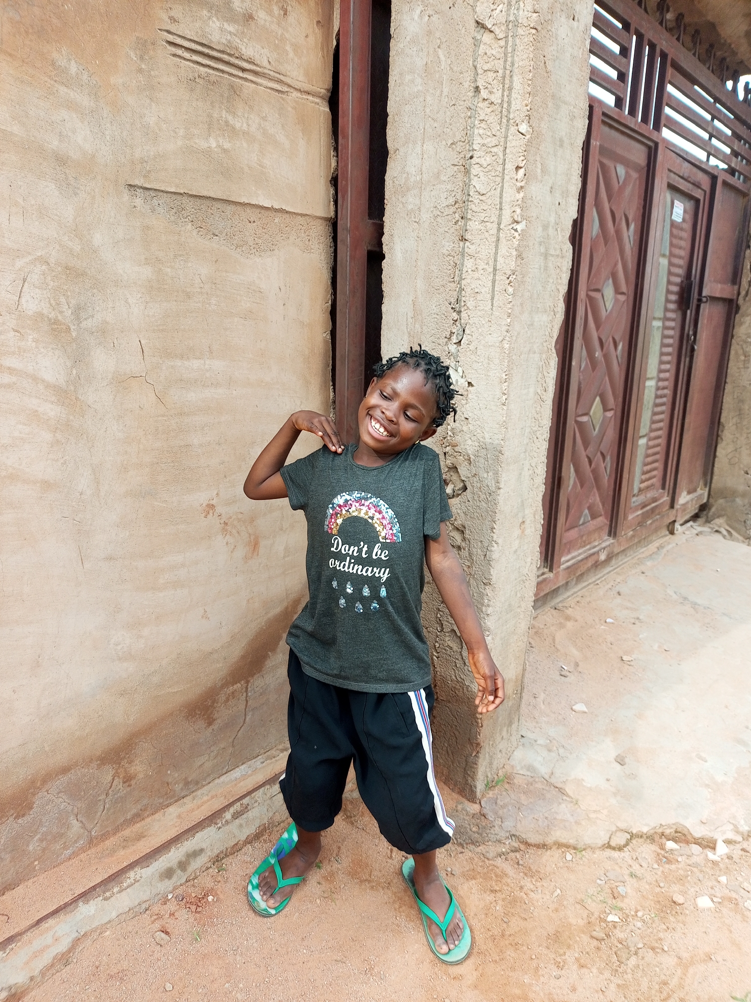 A young Black girl with braided hair is standing outside a building in Tempure, Nigeria. She is wearing a dark grey t-shirt with a rainbow graphic and text that reads "Don't be ordinary," and black shorts with a white stripe. She is smiling broadly and has her right arm raised, with her hand resting on her shoulder. The girl is wearing green flip-flops. She is standing in front of a tan-colored wall with some cracks and marks. To her right, there is a dark brown metal gate. The ground is covered in reddish-brown dirt and small stones.