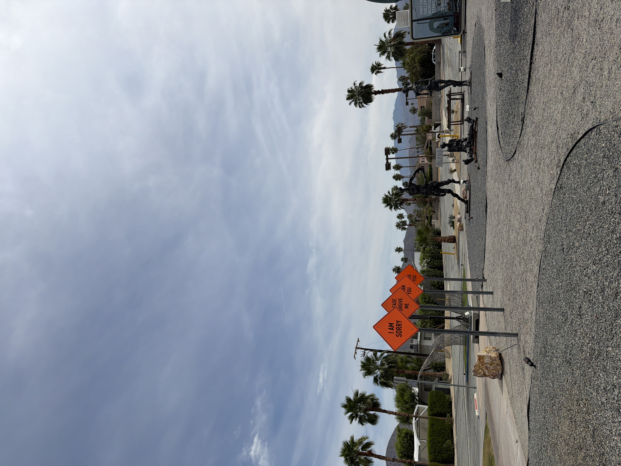 The image shows a street scene in Palm Desert, California, United States, on a sunny day with a mostly cloudy sky. Along the street are a series of four orange diamond-shaped signs reading "I AM SORRY", "EASE FORGIVE ME", "THANK YOU," and "HAVE YOU" respectively. The signs are mounted on metal posts. In the background are palm trees, mountains, and buildings, and along the sidewalk are black sculptures and gravel in curved patterns. A sign says "VISIT OL GALLERY ACROSS STREET" and "ALPOD" and "73-660 EL PA".