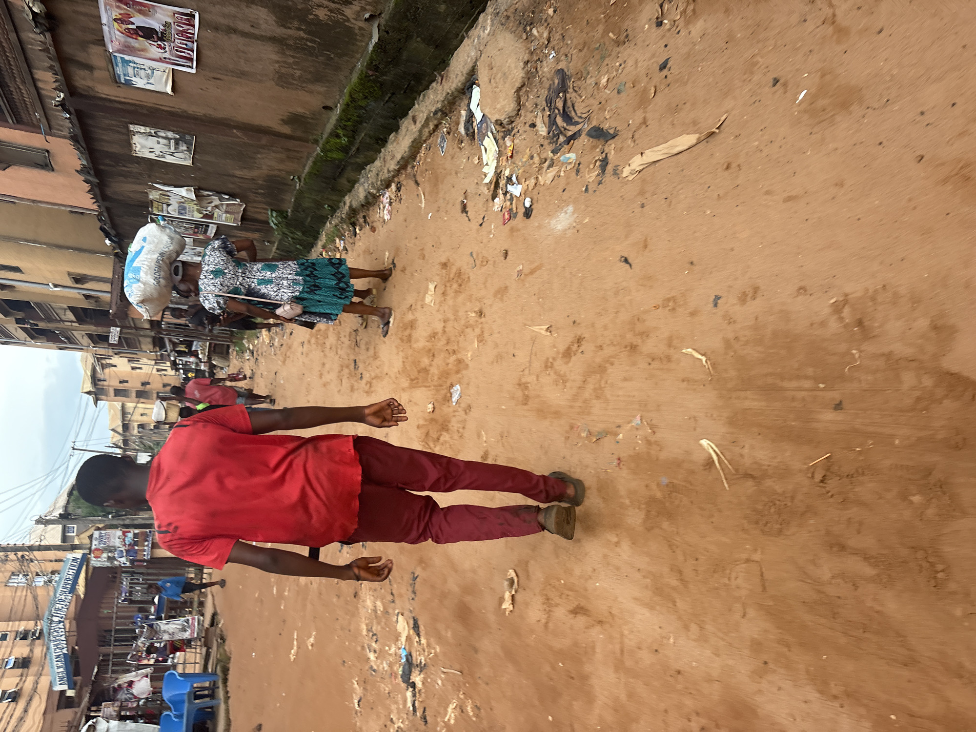 The image captures a street scene in Nkpor, Nigeria. In the foreground, a young Black male wearing a red t-shirt and maroon trousers walks away from the camera down a dirt road. Behind him, another Black female, also walking away from the camera, carries a large white bag of what appears to be sugar on her head. She is wearing a patterned dress and a cap. The street is unpaved and covered in reddish-brown dirt, with scattered debris and litter along the sides. Buildings line the right side of the street, with posters and advertisements affixed to a wall. On the left, there are blue plastic chairs outside a building with signage that reads "ANGLICAN CHURCH OF THE RESURRECTION." The overall environment appears to be an urban setting, possibly a market or residential area, characterized by its gritty, lived-in aesthetic. The sky is overcast.