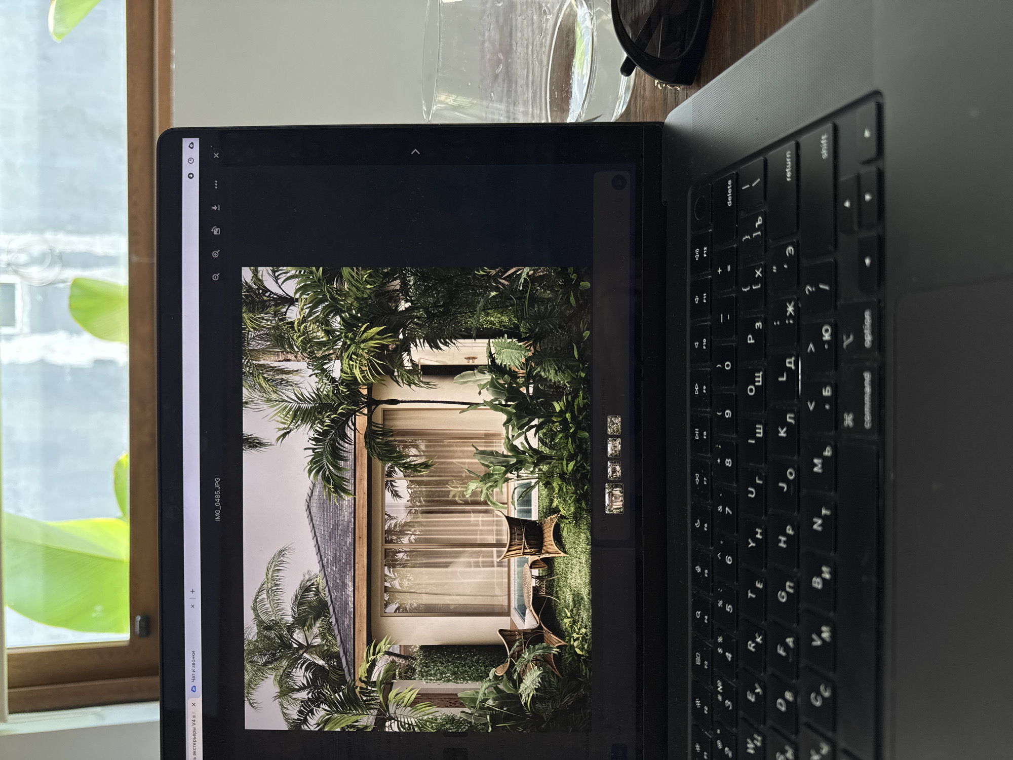 The image is a close-up shot of a laptop displaying a photo of an exterior scene, possibly of a residence or a resort, in a tropical location. The photo shows a building with a tiled roof and large windows, surrounded by lush greenery including palm trees and other tropical plants. There are chairs and a table on a small patch of grass, suggesting a patio area. The keyboard of the laptop is visible, and it appears to be a Cyrillic keyboard. Also on the table is a glass of water and a pair of sunglasses. The laptop is placed near a window, with a view of more greenery outside. The image does not contain people. While the description mentions a tropical location, the city of Denpasar, Indonesia, is not explicitly present within the visual details of the image itself.