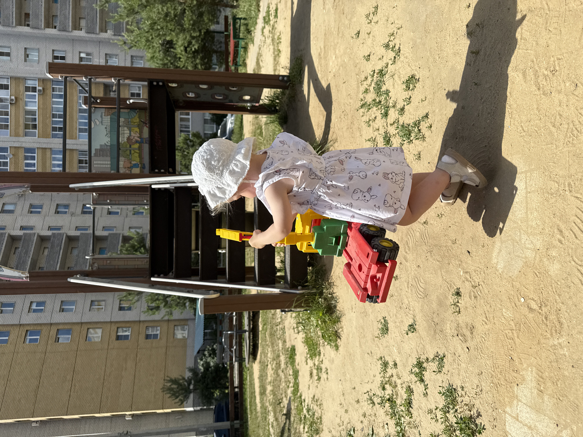 A toddler, facing away from the camera and wearing a white sun hat and a white dress with a bunny print, stands on a sandy playground. The child is holding a red and yellow toy truck with a green crane attachment. The playground is situated in front of a multi-story apartment building. The building's facade is a mix of yellow brick and grey concrete, with numerous windows. Trees and other playground equipment are visible in the background. The ground is covered in sand and some sparse green vegetation. The lighting suggests a sunny day, casting a distinct shadow from the child and the toy. The scene appears to be in a residential area of the city of Vladimir, Russia.