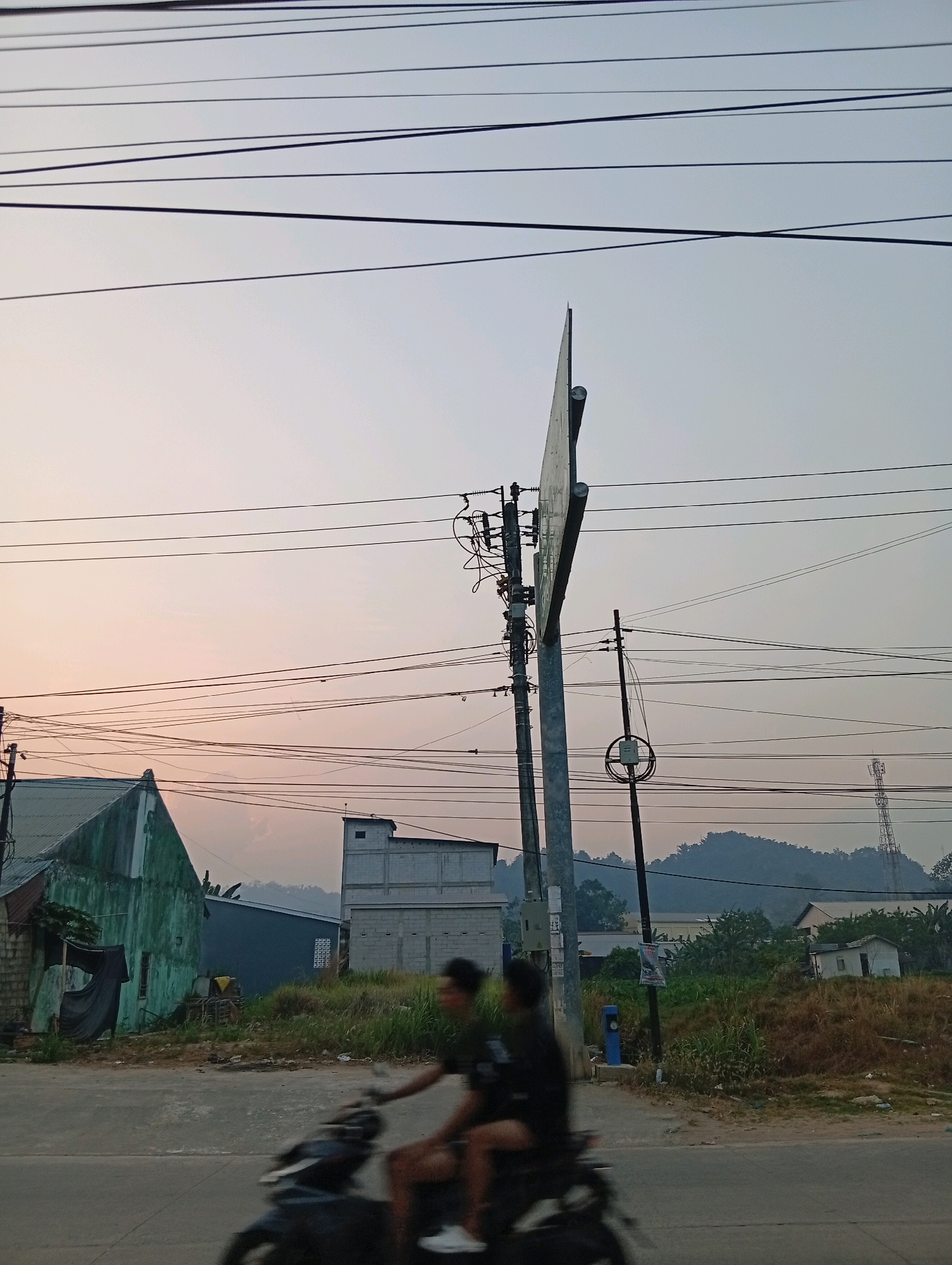 This is a photograph taken in Samarinda, Indonesia, showing a street scene at dusk. The sky is a soft gradient of pale orange and gray, suggesting the sun is setting. Overhead, a complex network of electrical wires crisscrosses the frame, casting faint shadows.
In the foreground, a motorcycle is captured in motion, its two riders blurred by the speed. The rider in the front appears to be wearing a dark t-shirt, and the passenger behind them is also in dark clothing. The motorcycle itself is dark-colored.
To the left of the road, there's a distressed, green-painted building with a corrugated metal roof. A dark fabric is draped over a window or opening. Adjacent to it are more modern-looking buildings, one gray and multi-story with visible windows, and another with a lighter facade. The ground is covered in dry grass and some scrub vegetation, with scattered debris.
A tall utility pole with various electrical components and wires stands prominently in the middle ground. Further in the background, a telecommunications tower is visible on a hill, alongside more buildings and trees. The overall impression is one of a semi-urban environment experiencing the transition from day to night.
