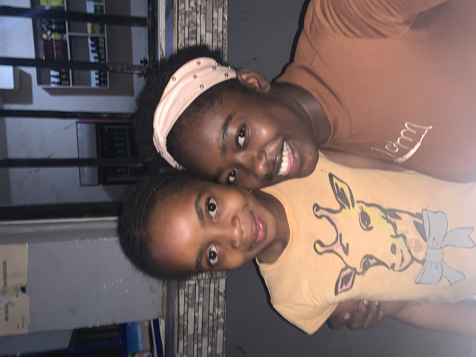 Two Black females are posing for a photo. The younger female is wearing a yellow t-shirt with a giraffe head on it. The older female has a beige headband with beads on it. The older female's t-shirt is brown and has white cursive writing on it. Behind them, there is a building in New Stadium, Nigeria. The building has windows and shelves with items on them.