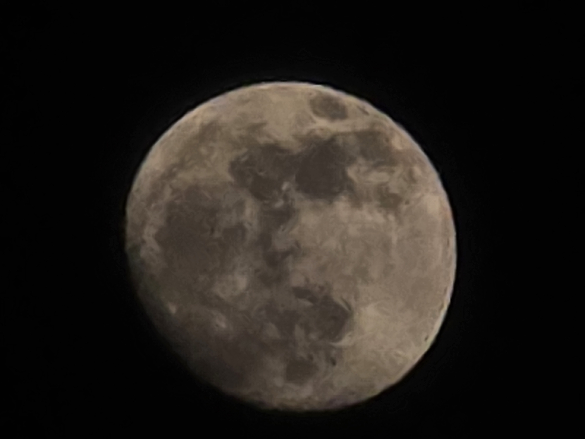 The image is a close-up, slightly out-of-focus photograph of the moon against a black, starless sky. The moon is large in the frame, taking up most of the space. Its surface is textured with variations in light and shadow, suggesting craters and maria (dark, flat plains). The colors are muted, ranging from pale beige to grayish-brown. There are no discernible features of the city of Yambol, Bulgaria, nor any people, specific objects, or actions present in the image. The scene is purely celestial, depicting the moon in its isolation.