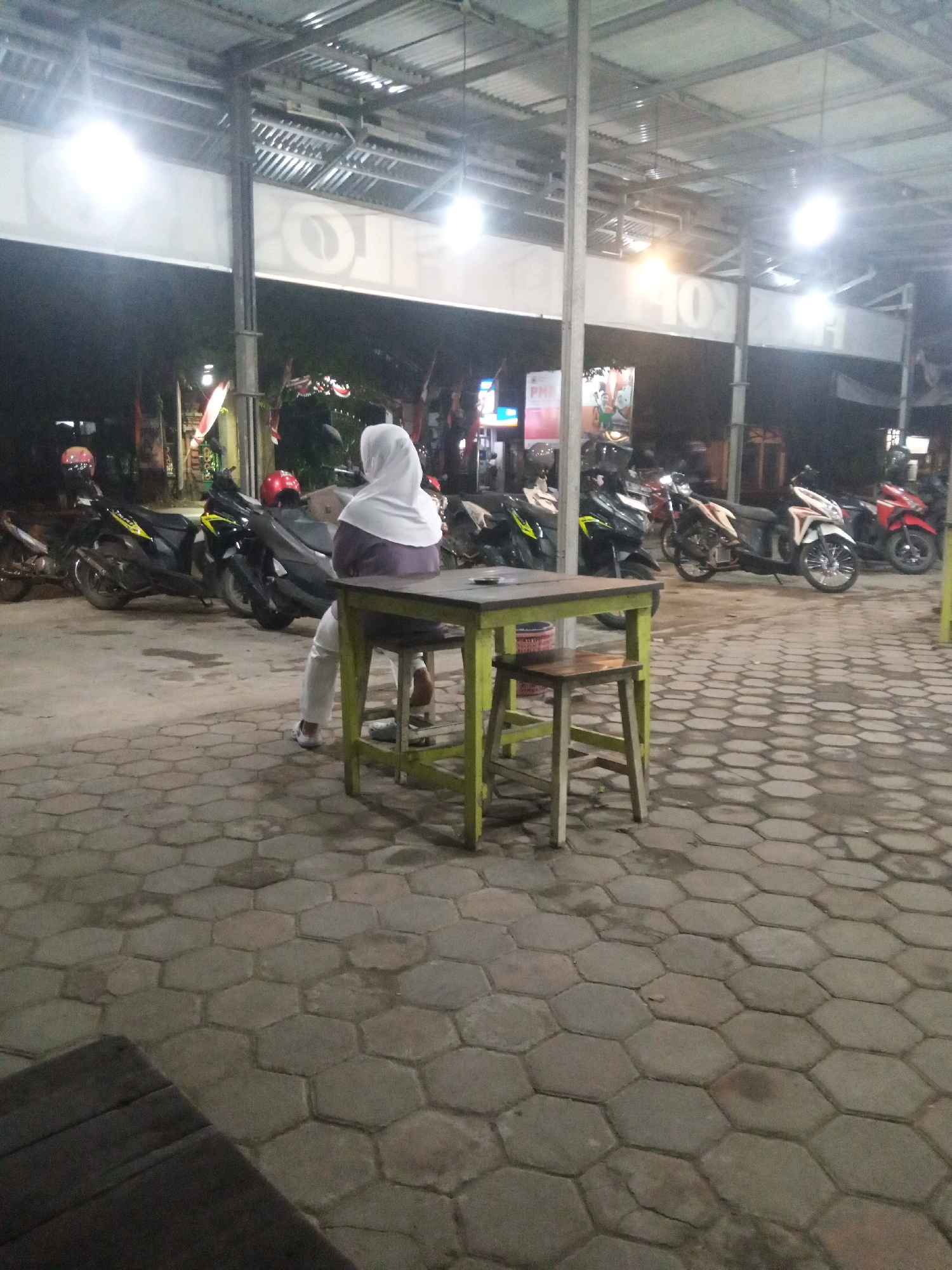A woman wearing a white hijab and a purple top sits on a stool at a green table on a hexagonal paved patio. She is facing away from the camera. Several motorcycles are parked behind her. The scene is outdoors, likely at night, with overhead lighting. A portion of a wooden table or bench is visible in the lower left corner of the image. The environment suggests a roadside eatery or gathering place in Gayaman, Indonesia.