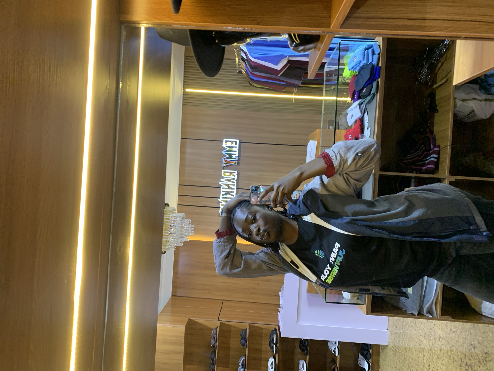 In Gura Top, Nigeria, a young woman takes a mirror selfie in what appears to be a retail establishment. She is wearing a dark t-shirt with a colorful logo that reads "PAIN YOUR UNIVERSE" in reverse, and an open grey jacket with red accents on the sleeves. Her hair is braided, and she is posing with one hand behind her head and the other holding a mobile phone.
The background reveals a well-lit interior with wooden shelving units filled with various items, including sandals on the left and what seem to be hats and folded clothing on shelves to the right. A prominent neon sign spelling "EMMY RANKIN" in reverse is visible on the back wall, reflecting in the mirror. A crystal chandelier hangs from the ceiling above the sign. To the right, a mannequin is partially visible, wearing a black hat. The overall environment suggests a fashion boutique or a similar retail space.