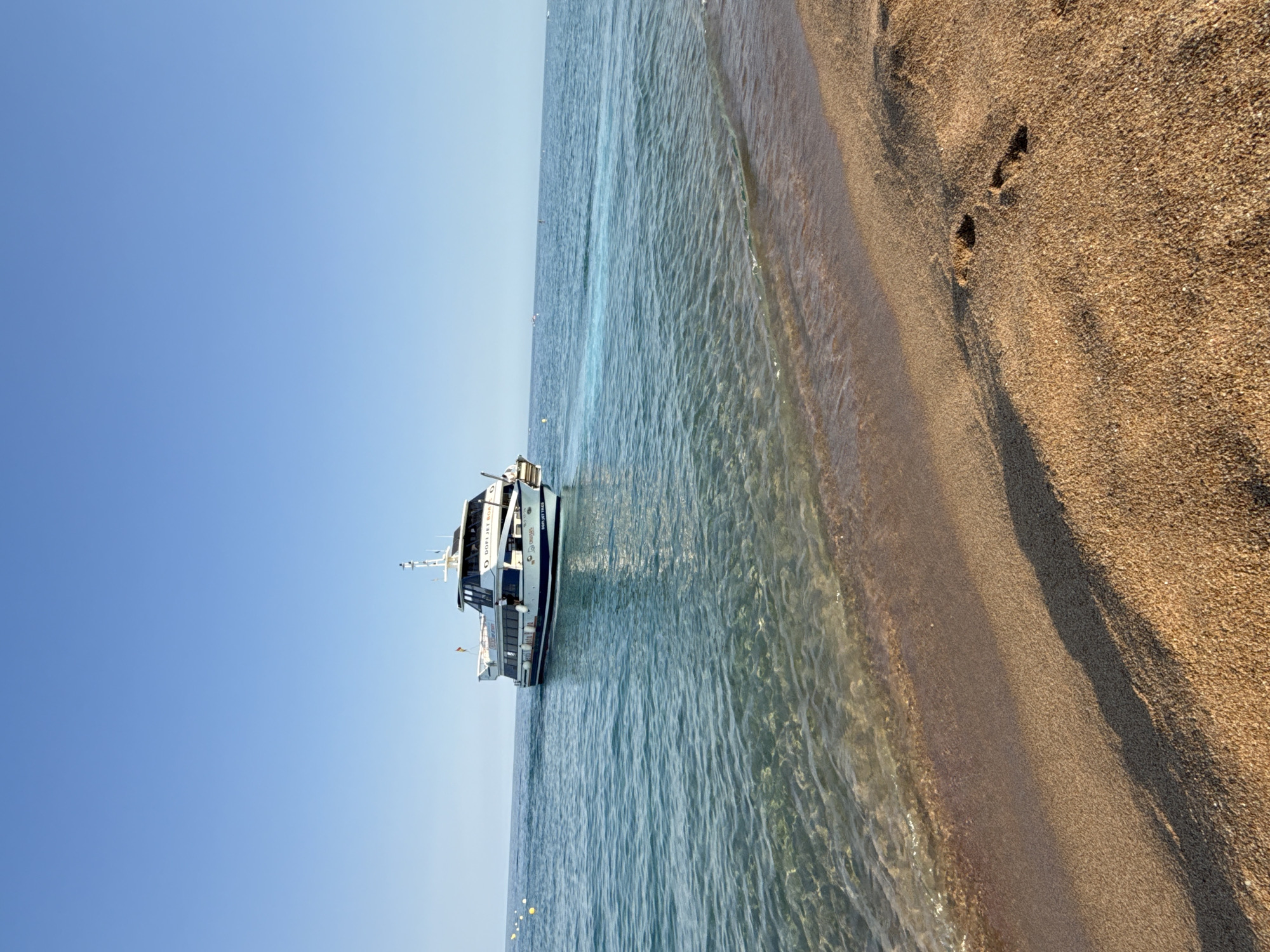 The image depicts a sunny day at a beach in Blanes, Spain. The foreground is dominated by golden sand with a few visible footprints. The water is clear and calm, with gentle waves lapping at the shore. In the mid-ground, a white passenger boat with blue trim is cruising on the sea, leaving a noticeable white wake. The boat has "DOFI JET BOAT" written on its side. The background shows a clear, bright blue sky with no clouds. The overall scene conveys a peaceful and inviting coastal environment.