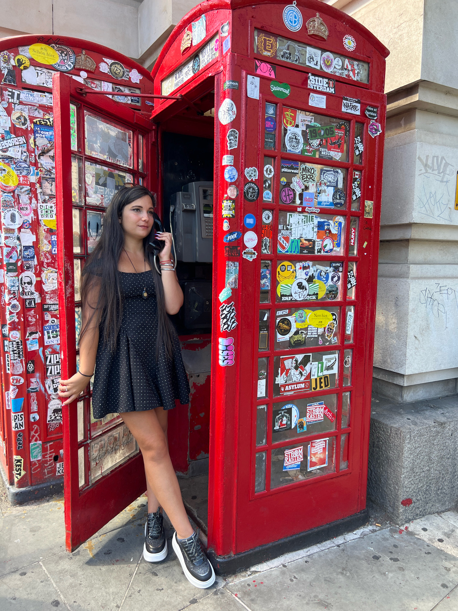 A young woman with long, dark hair is standing in an open red telephone booth, holding a black telephone receiver to her ear. She is wearing a black, polka-dotted dress and black and white sneakers. The telephone booth is covered in stickers, and another red telephone booth is visible to her left, also covered in stickers. The booths are situated on a sidewalk next to a stone building. The scene appears to be in London, United Kingdom.