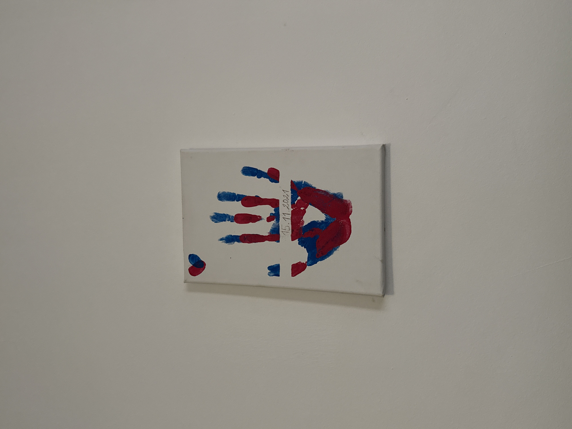 A child's canvas painting, painted in red and blue, is mounted on a light-grey wall. The painting appears to be a handprint, with the palm and thumb rendered in red, and the fingers in blue. A red and blue heart is located in the lower left-hand corner. The date "15.11.2021" is written vertically in black ink over the center. Location: Unknown, Unknown.