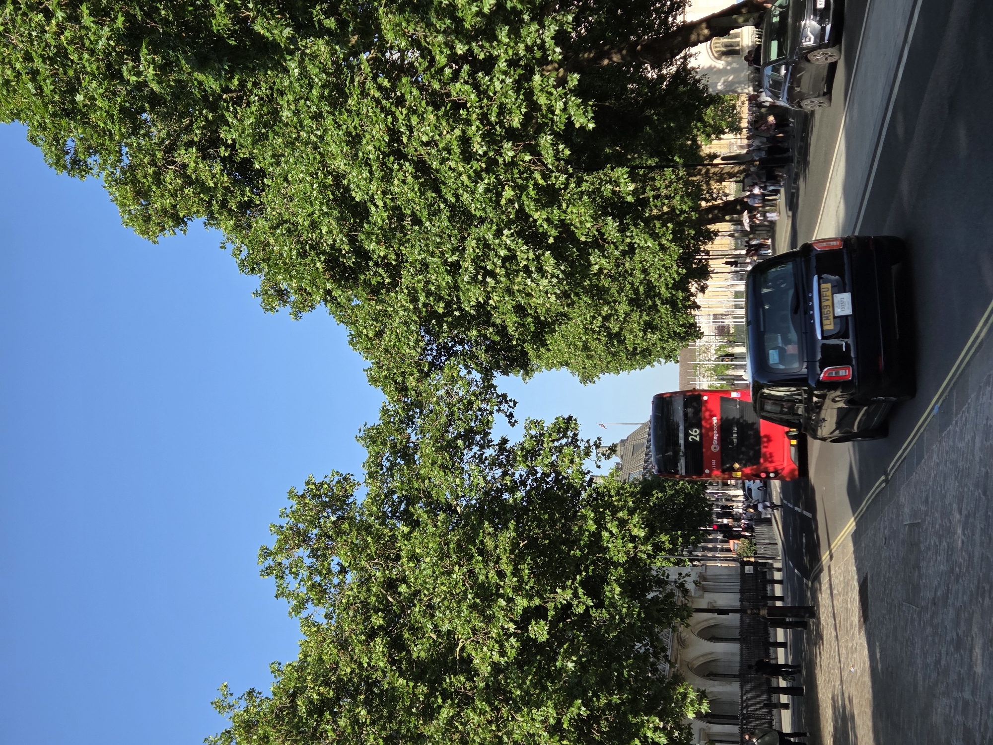 This image shows a street in London, United Kingdom. Two rows of trees frame the blue sky. The street contains a black taxi, a red double-decker bus labeled "26," and other vehicles. On the sidewalk, there are pedestrians and a fence bordering a building. The architecture of the building indicates a location in the city of London.