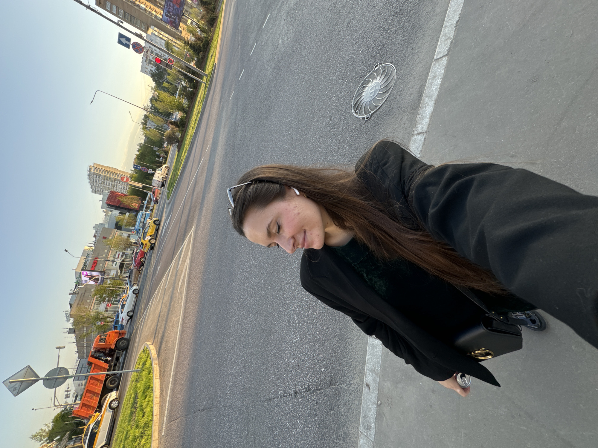 A woman is taking a selfie while standing on a sidewalk in Moscow, Russia. The woman has long brown hair and is wearing a black jacket. She has sunglasses on her head and is carrying a black purse. In the background is a street with cars, buses, and buildings. The sky is clear and blue. An orange dump truck is also visible.