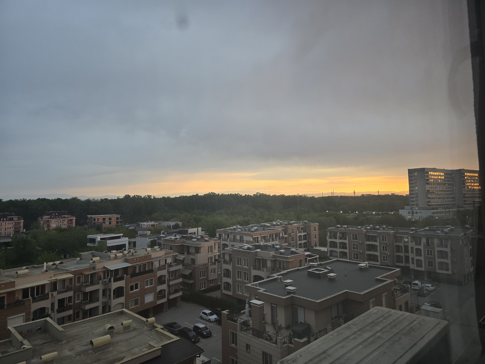The image displays an elevated view of a residential area during either sunset or sunrise. The sky is predominantly covered by a thick layer of grey clouds, but a distinct band of vibrant orange and yellow light is visible along the horizon, indicating the sun's position.
The foreground and midground are dominated by numerous multi-story apartment buildings, characterized by their contemporary architecture, balconies, and flat roofs adorned with various fixtures like ventilation units. The buildings exhibit a palette of neutral colors, including beige, brown, and grey. Several parked cars are visible in the parking lots and courtyards nestled between these structures.
A dense, dark green expanse of trees stretches across the middle ground, separating the immediate residential blocks from the distant horizon. On the far right, a tall, modern building with many windows reflects the golden light from the horizon. Faint outlines of mountains or hills can be discerned on the far left, beyond the tree line. A dark window frame partially obscures the right edge of the photograph, suggesting the image was captured from inside a building.
The scene is devoid of people. No discernible text is visible anywhere in the image. The overall impression is one of a quiet urban or suburban landscape bathed in the soft, fading or rising light, under an overcast sky.