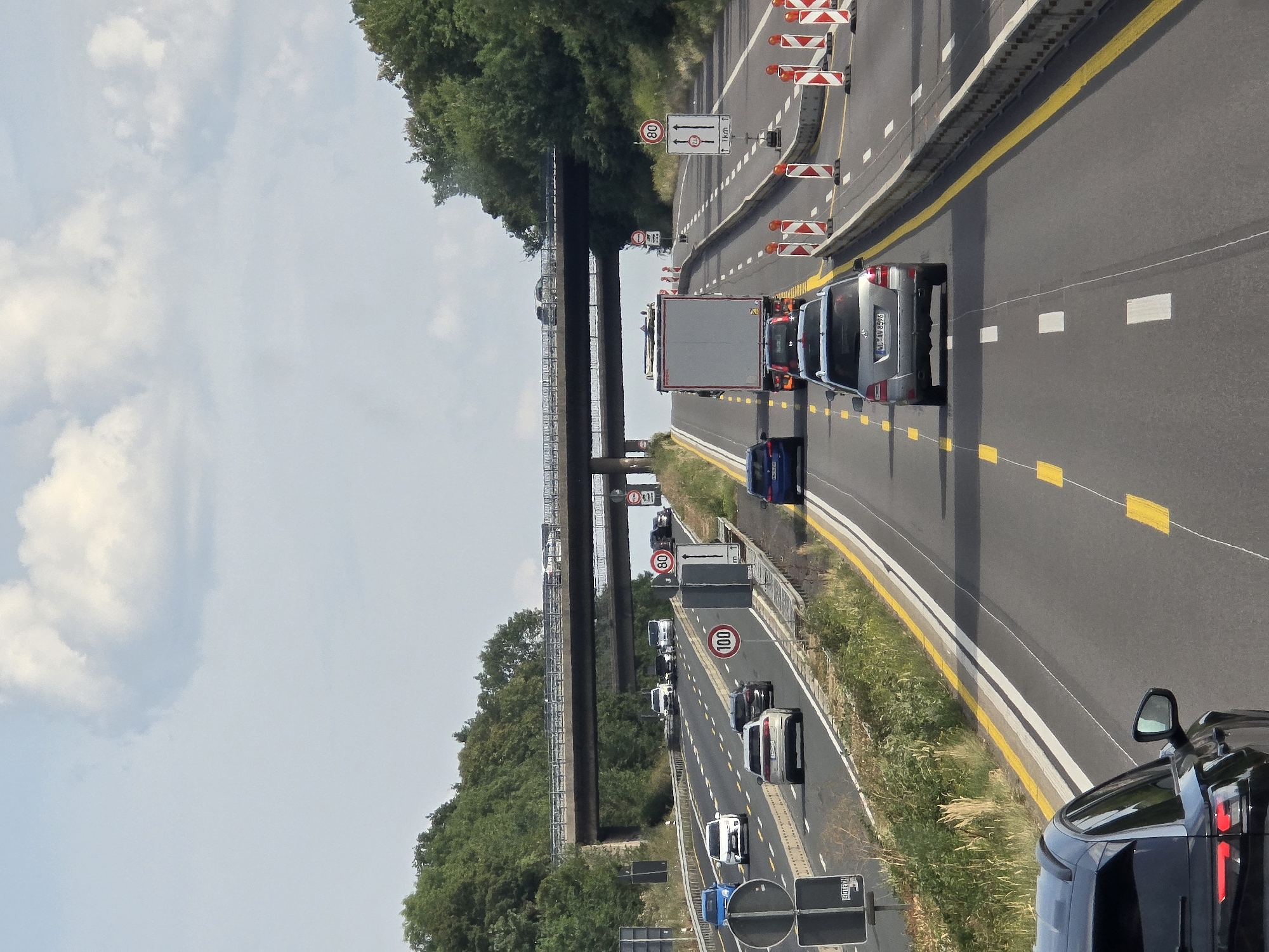 This is a vertical, eye-level, full shot of a multi-lane highway in Königswinter, Germany, under a bright, cloudy sky. The highway has two visible directions of travel, separated by a concrete barrier and dotted yellow and white lines. In the foreground, a dark colored SUV is in the right lane. Further ahead, a silver Mercedes-Benz sedan is in the center lane, followed by a white semi-truck and another silver sedan. Traffic is light to moderate in this direction, with a few other vehicles visible in the distance. The direction of travel going away from the viewer has more cars, including a blue sedan, a white sedan, and a van.
On the right side of the highway, there is a construction zone with orange and white traffic cones and barriers. Several speed limit signs are visible: one for 80 km/h and another for 100 km/h. An overpass carrying another section of highway is visible in the background, with traffic also visible on it. Trees line both sides of the highway, with a dense forest covering the hillsides. The overall scene depicts a typical German highway during the daytime, with some road construction occurring.