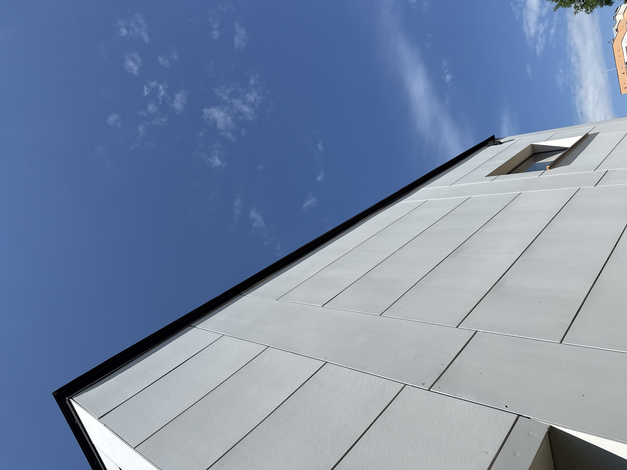 The image depicts the modern facade of a building, characterized by light gray, horizontal paneling with thin vertical seams. A dark, angled roofline runs along the top left of the structure. A small, rectangular window is visible on the right side of the building, recessed within the wall. The building is set against a clear, vibrant blue sky with scattered wispy white clouds. In the lower right corner, the edge of another building with an orange facade and a tree branch are partially visible. The scene is shot from a low angle, emphasizing the architectural lines of the building and the expanse of the sky. The location is specified as Sofia, Bulgaria.