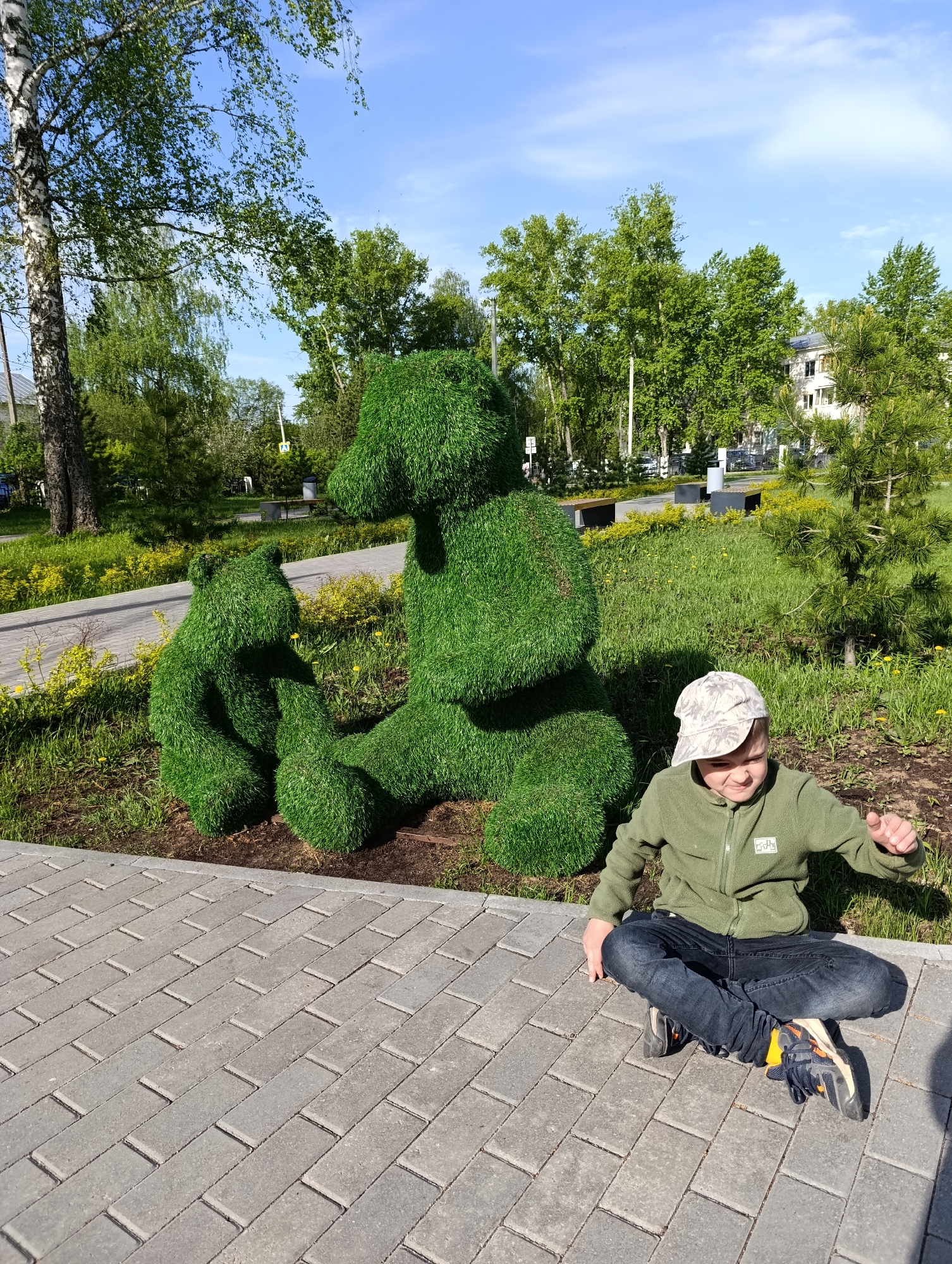 The image captures a young boy seated cross-legged on a gray brick walkway, positioned in front of a pair of topiary bear sculptures in Chaikovsky, Russia. The boy is wearing a green fleece jacket, dark jeans, and a patterned baseball cap. The bear sculptures, appearing to be made of dense green foliage, consist of a larger bear sitting upright next to a smaller, seated bear cub. The background includes a grassy area with various trees and shrubs, as well as parts of buildings under a partly cloudy sky.