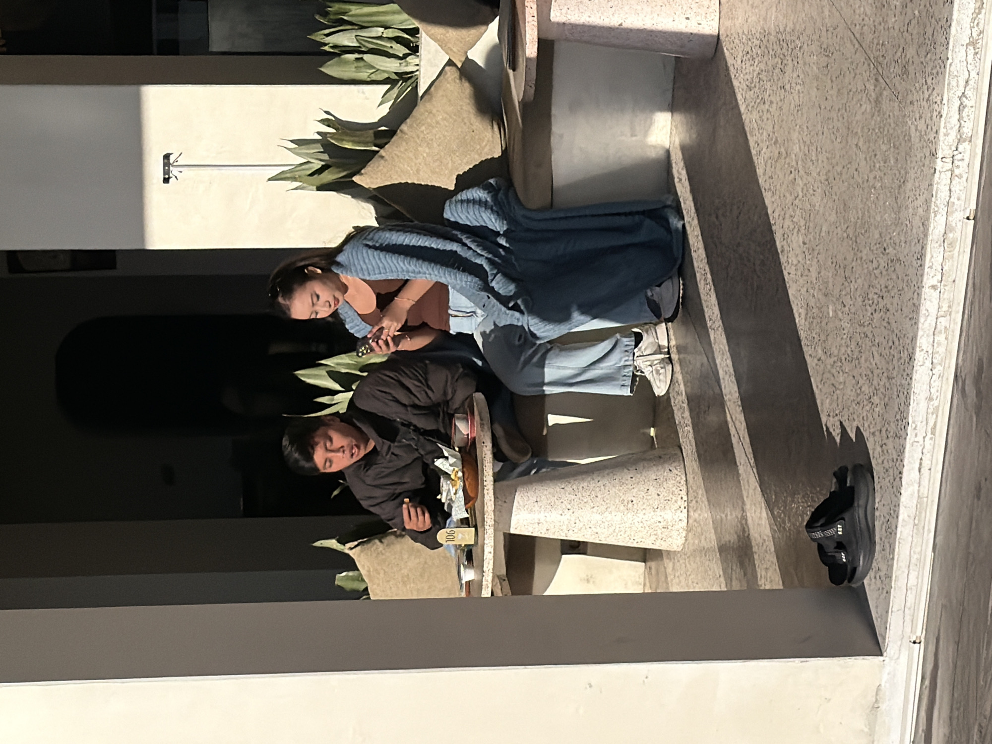 The image depicts two individuals, a male and a female, seated outdoors at a cafe or restaurant in Kintamani, Indonesia, under strong, low-angle natural light that casts prominent shadows.
The male, positioned on the left, appears to be in his late teens or early twenties, dressed in a dark, possibly padded jacket and blue jeans. He is actively eating from a wooden bowl on the table, holding food to his mouth with his right hand, his mouth slightly open.
The female, also appearing to be in her late teens or early twenties, is seated to the right of the male. She is wearing a brown tank top, light blue wide-leg jeans, and white sneakers, and is wrapped in a large blue knitted blanket. She is holding a smartphone in both hands and looking down at its screen, seemingly absorbed in its content. Her dark hair is pulled back.
Between the two individuals is a light-colored, possibly terrazzo, round table with a conical base. On the table are a wooden bowl containing what appears to be fried food (such as french fries) wrapped in paper, a small white condiment cup, and a yellow table number sign displaying "106." A dark cup or bowl is also present near the female.
Both individuals are seated on a built-in bench with light brown, patterned cushions. Behind them, along the back wall, are multiple green, leafy plants. A vertical pole with a small black device, possibly a security camera or sensor, is mounted on the wall behind the female. To the far right, a portion of another smaller, light-colored round table is visible.
In the foreground, near a white vertical pillar, a pair of black athletic sandals are casually placed on the light gray, speckled concrete floor. The floor shows distinct, elongated dark shadows cast by the pillars and possibly the seated figures, indicating the strong directional sunlight. The scene is framed by white and dark gray architectural elements.
