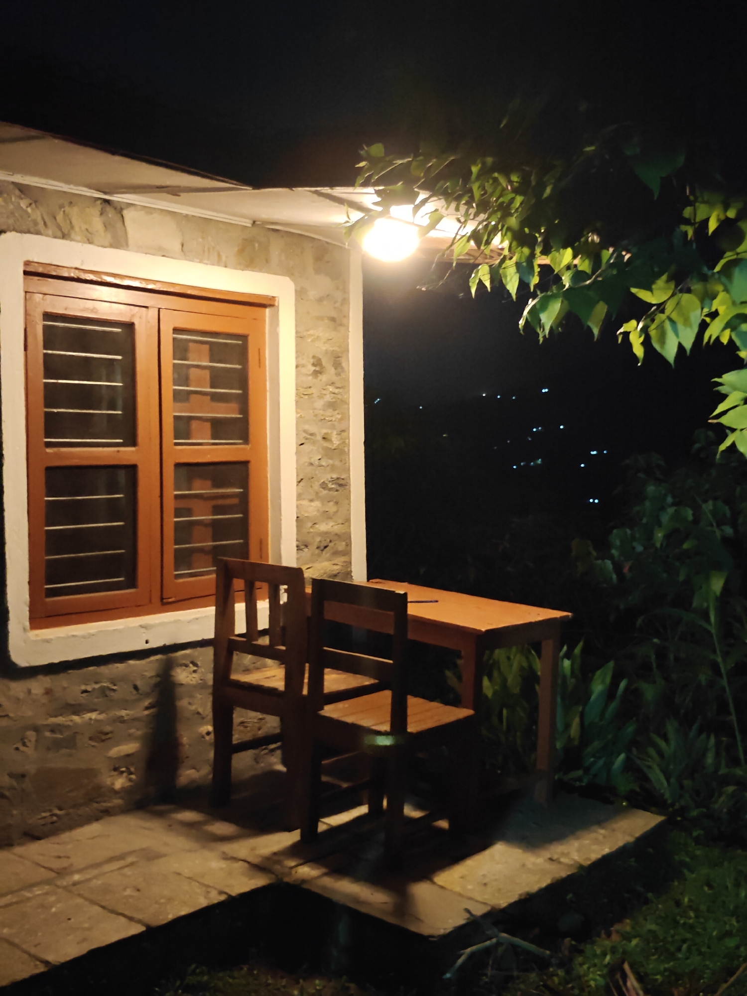 A wooden table and two chairs sit on a paved patio in front of a stone-walled building with a wooden-framed window at night. A single light bulb hangs from the ceiling, illuminating the scene. In the background, distant lights twinkle in the darkness, suggesting the presence of a city or settlement on a hillside. Lush green foliage frames the scene on the right, with dark trees and bushes surrounding the patio area. The overall atmosphere is one of quiet solitude and natural beauty.