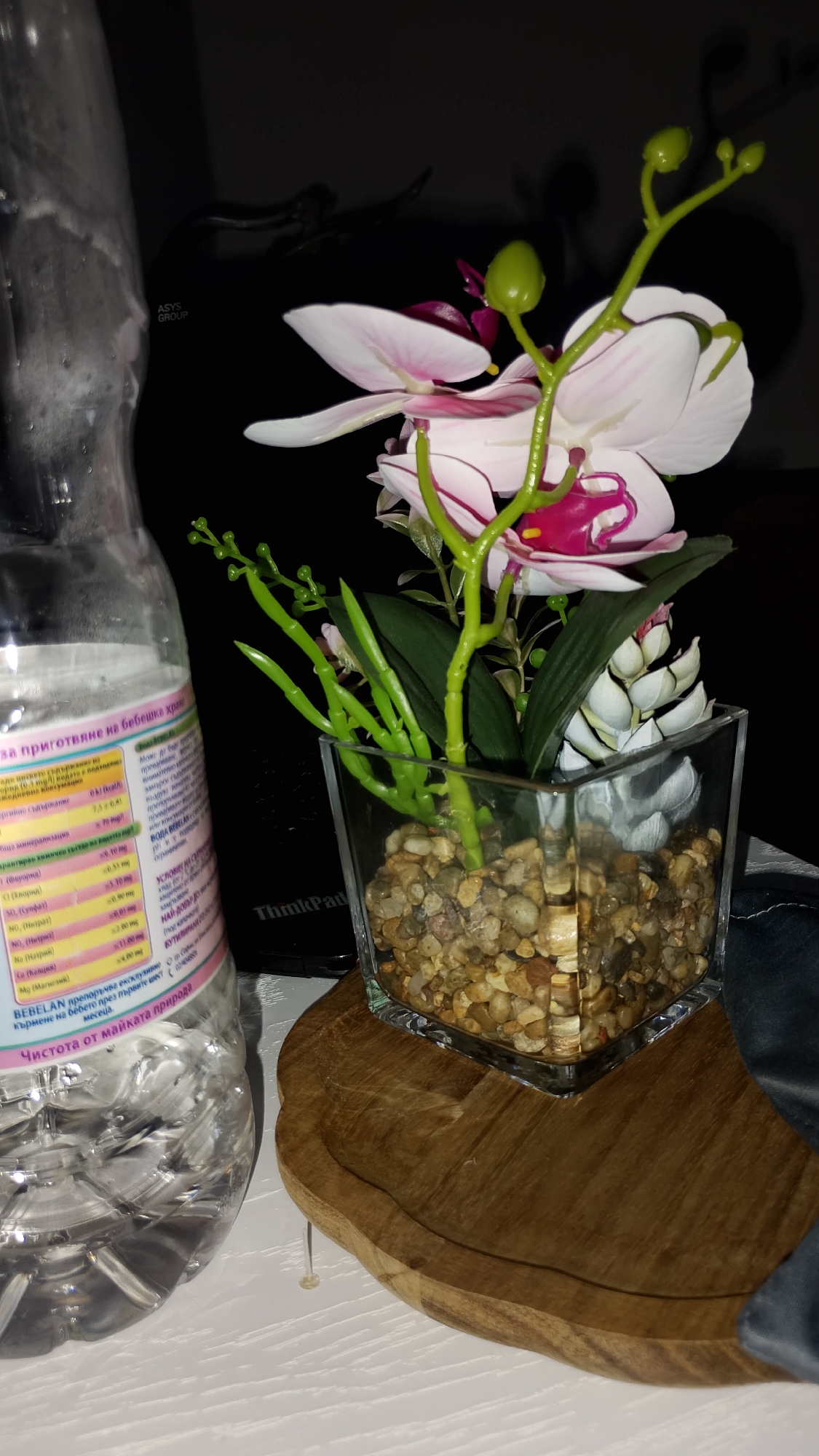 A clear plastic bottle of water sits next to a small glass vase filled with pebbles, in which a decorative orchid plant is placed. The orchid has pink and white petals and green stems with buds. A dark, possibly wooden, coaster-like object is positioned below the vase. In the background, a black electronic device, possibly a laptop, is visible with the "ThinkPad" logo. The lighting is dim, creating shadows. The image does not contain any specific information about the city of Smolyan, Bulgaria.