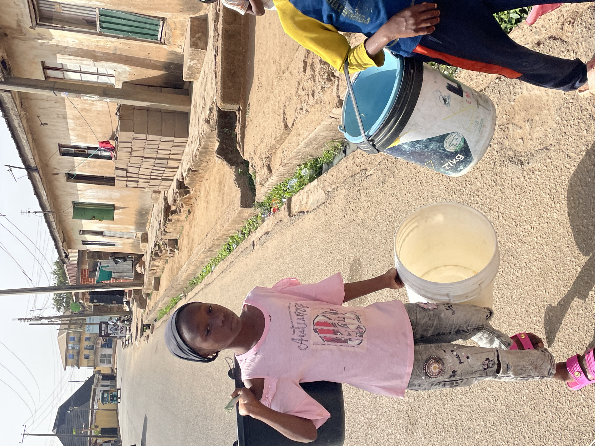 This image, captured in Jos, Nigeria, presents a street scene under bright daylight, slightly rotated to the left. The ground is a mix of uneven dirt and concrete, with a narrow, vegetation-filled gutter running alongside.
In the foreground, a young girl, estimated to be between 7-10 years old, stands looking directly towards the viewer. She is dressed in a light pink short-sleeved t-shirt with a stylized graphic including the word "Autumn," grey patterned trousers, and bright pink sandals. She holds a white, empty plastic bucket with her right hand and appears to be clutching green paper currency in her left.
Behind her, another individual, likely also a young person, is partially visible, wearing a blue long-sleeved top with yellow sleeves and dark trousers featuring an orange stripe. This person carries a blue plastic bucket that is partially filled with water. Visible text on the blue bucket includes "21kg" and "Afrika," along with some warning symbols and other partially obscured lettering.
The background features a row of buildings constructed from light-colored, possibly unpainted concrete or stucco. Some buildings have windows with green shutters, and one structure displays a stack of grey rectangular blocks on a lower ledge. Power lines are visible against a clear, bright sky. Further down the street, more buildings and a sign reading "THE CELL POS" can be seen. The strong shadows suggest it is midday or early afternoon. The scene illustrates the activity of carrying water buckets, a common daily task in the region.