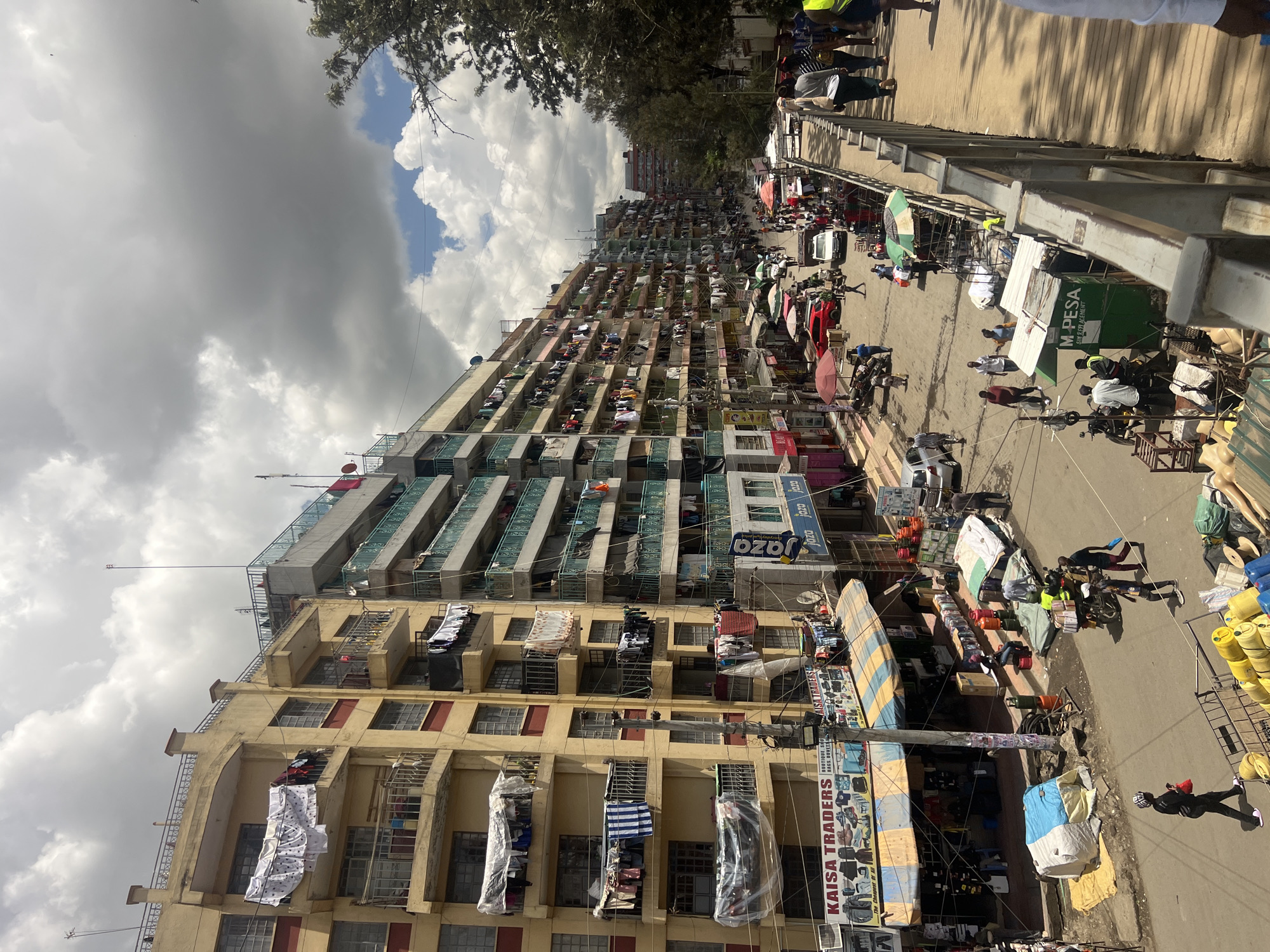 The image depicts a densely populated urban area, likely in Nairobi, Kenya. Tall, multi-story buildings line a street, with clothes hanging from the balconies, indicating residential use. The buildings are of varying architectural styles, some with weathered facades. The street is bustling with activity, with numerous people walking, trading, and going about their business. Street vendors and stalls line the street.
Signage for 'KAISA TRADERS', 'Jaza', and 'M-PESA' are visible. The sky is partly cloudy with a mix of dark storm clouds and patches of blue. A bridge or overpass runs along the top edge of the frame, with pedestrians visible upon it.
