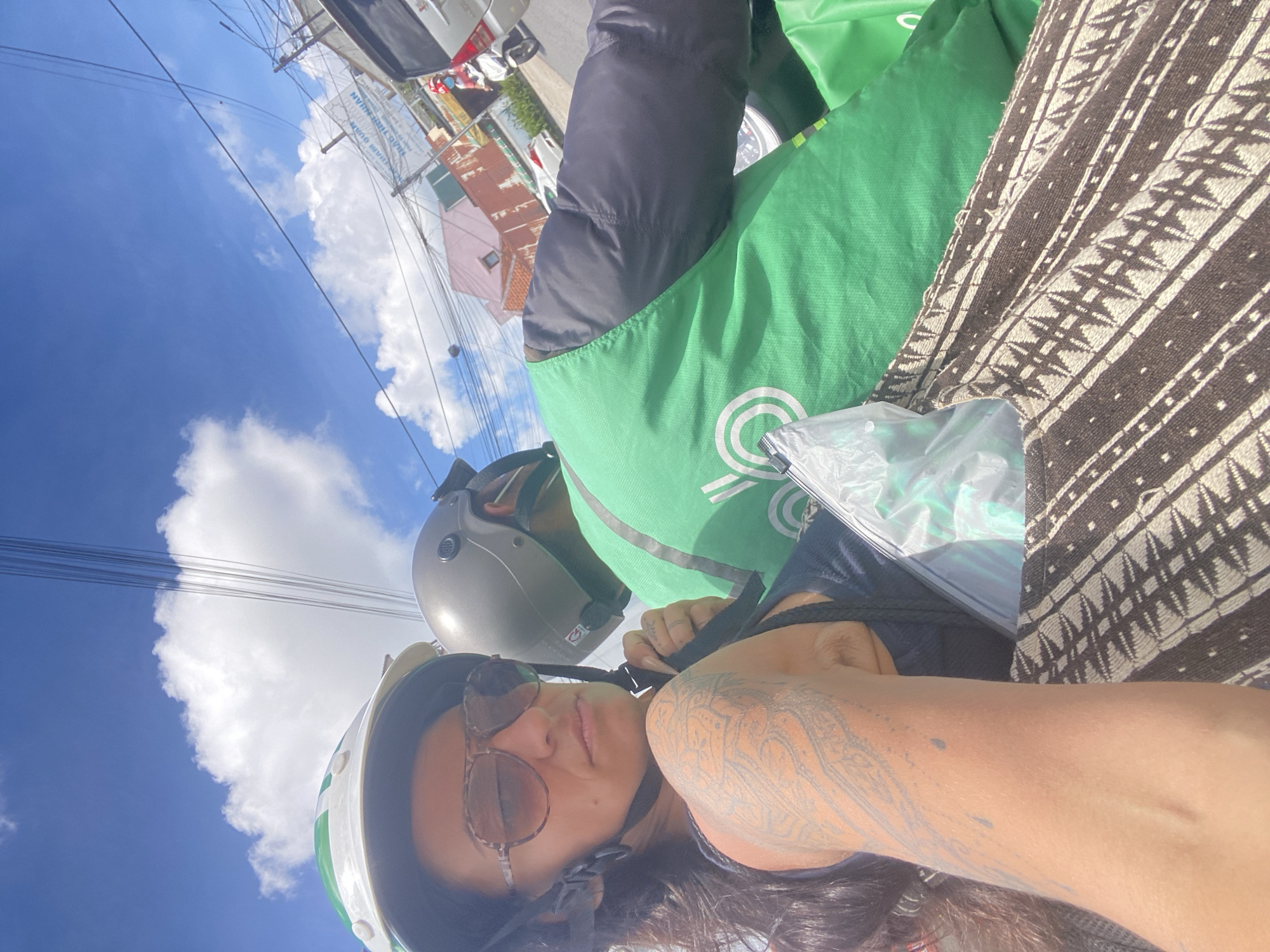 A woman with dark hair, sunglasses, and a helmet is riding on the back of a motorcycle, taking a selfie. She has a large tattoo on her arm. The motorcycle rider, wearing a green vest with a white logo and a gray helmet, is visible in front of her. They are on a street in Da Lat, Vietnam, with buildings and power lines visible in the background. The sky is blue with some clouds. A patterned bag is in the foreground.