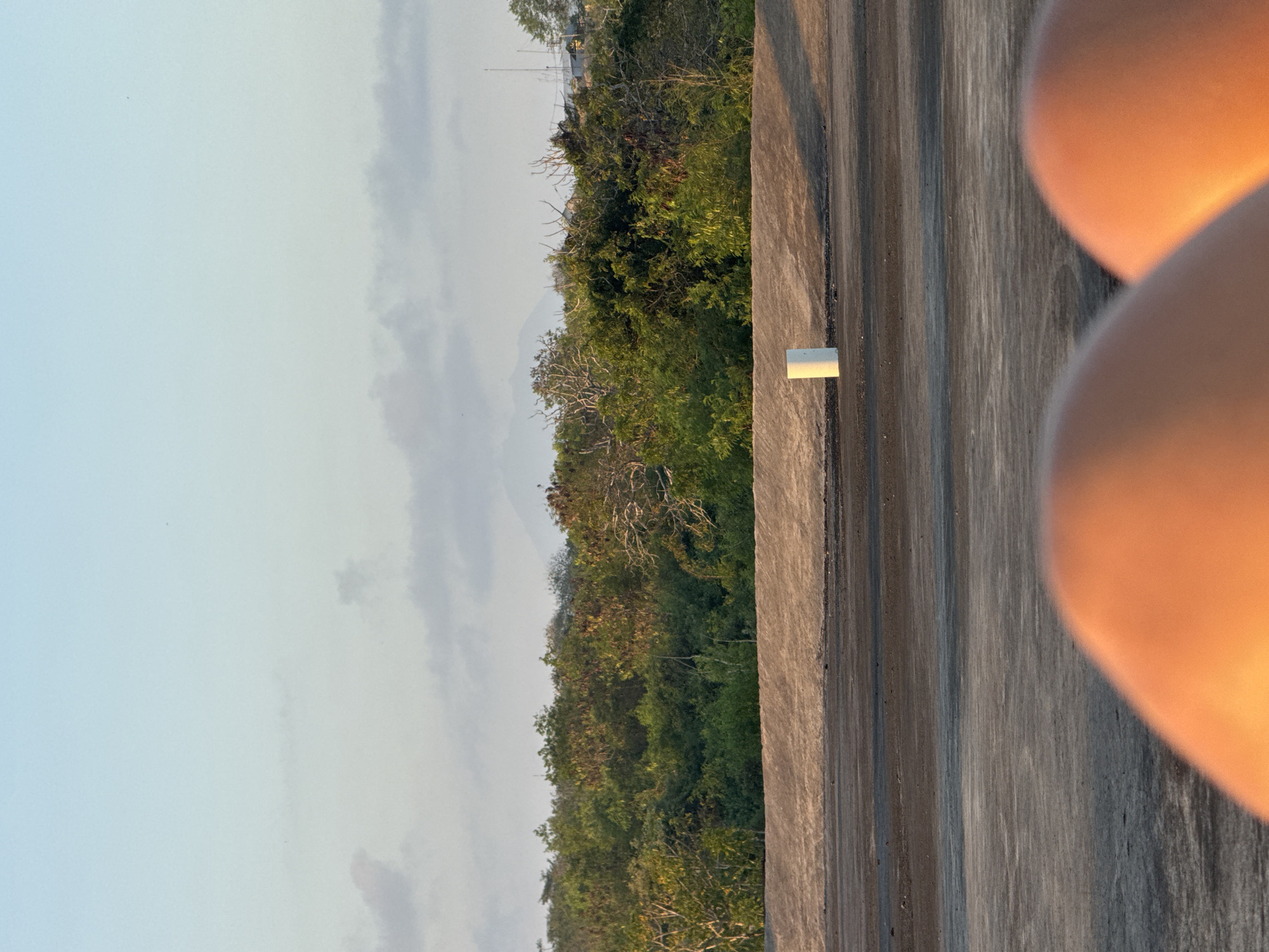 The image depicts an outdoor scene with a focus on a concrete structure in the foreground and a lush green landscape in the background. The concrete surface appears weathered and has distinct vertical lines, suggesting it might be a wall or a pathway. A small white cylindrical object is placed on the concrete surface. To the right, out of focus, are two large, softly rounded shapes that are orange-hued, possibly parts of a person's body or clothing illuminated by warm light.
The background reveals a dense collection of trees and vegetation, indicating a natural or semi-natural environment. In the distance, faint outlines of hills or mountains can be seen through a hazy sky. The sky itself is a pale blue with wispy clouds, suggesting it could be early morning or late afternoon. The overall lighting, particularly the warm glow on the out-of-focus shapes, points towards either sunrise or sunset.
The scene evokes a sense of tranquility and natural beauty, contrasted with the man-made concrete element. The presence of the softly blurred orange shapes suggests a human element is present, but their indistinctness adds an air of mystery or perhaps intimacy to the scene. There is no visible text in the image. Given the location context of Kuta Selatan, Indonesia, the dense vegetation could be characteristic of the region's tropical climate.