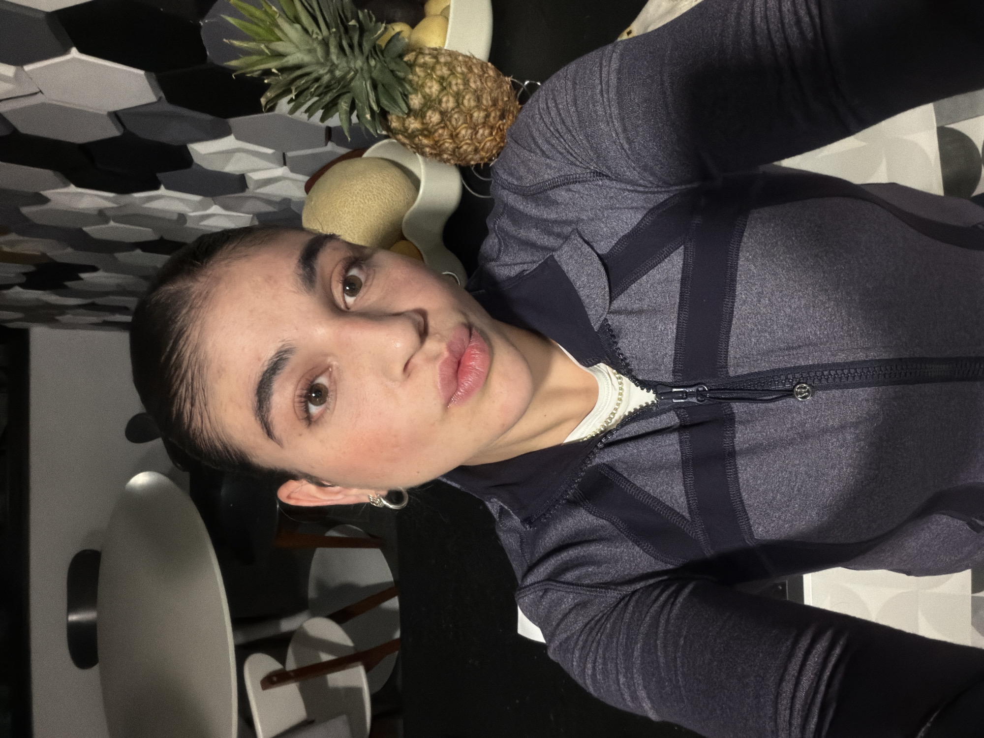 A selfie shows a young woman looking directly at the camera, with a neutral expression and slightly pouting lips. Her dark hair is pulled back, and she has light blush on her cheeks. She is wearing a dark grey athletic zip-up jacket with darker grey or black paneling, a white t-shirt, and a silver chain necklace. A small, silver circular logo, resembling the Lululemon emblem, is visible on the jacket's zipper pull.
The setting is indoors, likely a kitchen or dining area in Puebla, Mexico. To the right of the woman, a white fruit bowl is visible, containing a pineapple, a cantaloupe, and other assorted fruits. The fruit bowl rests on a dark countertop or table. Behind the fruit and to the left of the woman, there's a distinctive wall with a geometric pattern of black, grey, and white hexagonal tiles. Further back, parts of white chairs with wooden legs can be seen. The lighting is even and typical of an indoor environment. The image itself is rotated 90 degrees clockwise, presenting the scene tilted.