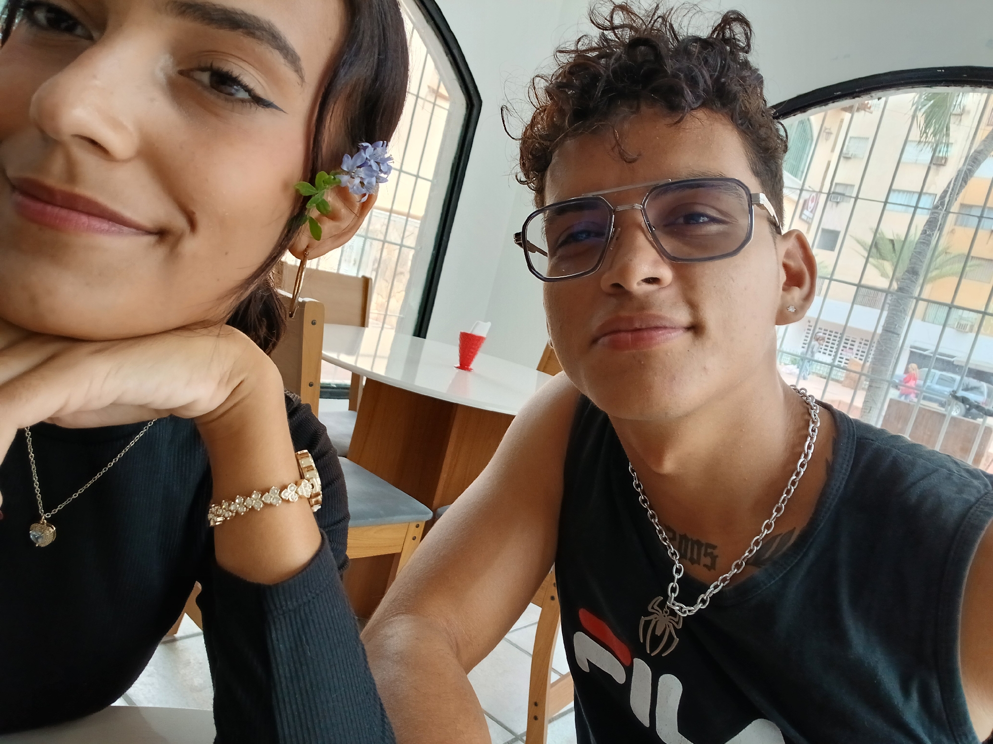 A close-up selfie shows two people indoors. On the left is a person with dark hair and brown eyes, wearing a black top, a necklace with a heart pendant, and a jeweled bracelet. They have a small blue flower behind their ear. On the right is a person with curly brown hair, wearing glasses, a black sleeveless shirt with a white and red logo, and a silver chain with a spider pendant. A visible tattoo is on their chest. They both look toward the camera.
Behind them is a white table with a red object on it, chairs, and a window. Outside the window, a multi-story building, trees, and a partial view of the street can be seen. Based on the architecture and vegetation, the location is likely within the city of Juan Griego, Venezuela.