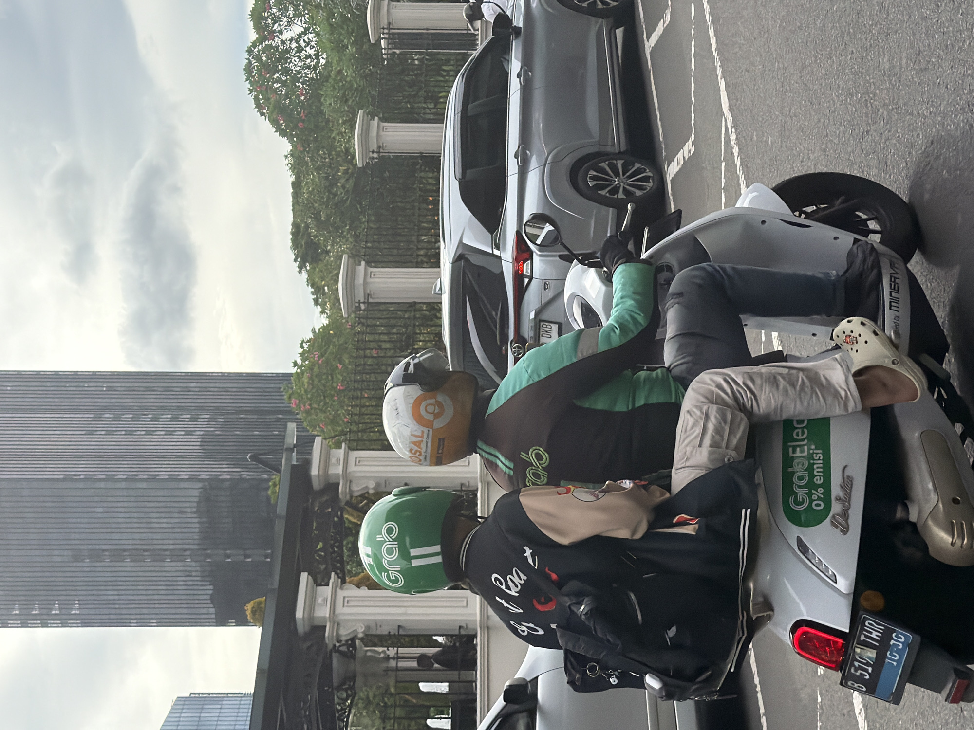 Two GrabBike drivers, wearing helmets, are waiting on their motorcycles on a city street in South Jakarta, Indonesia. One driver has a green helmet and the other has a white and brown helmet. Both drivers are wearing black jackets. The motorcycle in the foreground has a white body with a "GrabElec 0% emisi" sticker on the side. The license plate of the motorcycle reads "B 519 THR" and "10-30". A silver car is parked behind the motorcycles. Tall buildings and trees are visible in the background, along with a fence and white pillars.
