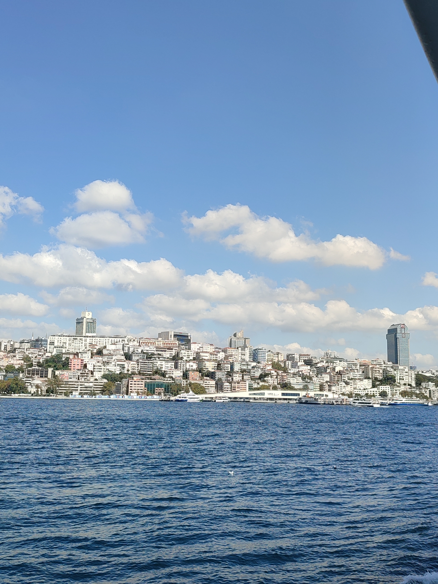 The image captures a panoramic view of the Beyoğlu district in Istanbul, Turkey, as seen from the Bosphorus Strait. The scene is dominated by the vibrant blue water in the foreground, with gentle ripples suggesting calm conditions. In the middle ground, a dense urban landscape unfolds, characterized by a multitude of buildings of varying heights and architectural styles, densely packed on a hilly terrain. White and pastel-colored structures intermingle with darker-toned buildings, creating a textured mosaic. Prominent among the cityscape are several notable landmarks: a tall, modern skyscraper with a distinctive blue facade stands out on the right side of the image, while a more traditional, elongated building with a prominent clock tower is visible further to the left. Smaller buildings with white facades and red-tiled roofs are scattered throughout the hillside, interspersed with patches of green trees. Several ferries and boats are docked or cruising along the waterfront, indicating active maritime transport. The background is filled with a bright blue sky, dotted with fluffy white cumulus clouds, suggesting a pleasant, sunny day. The overall impression is one of a bustling, historic, and scenic urban environment situated by the water.