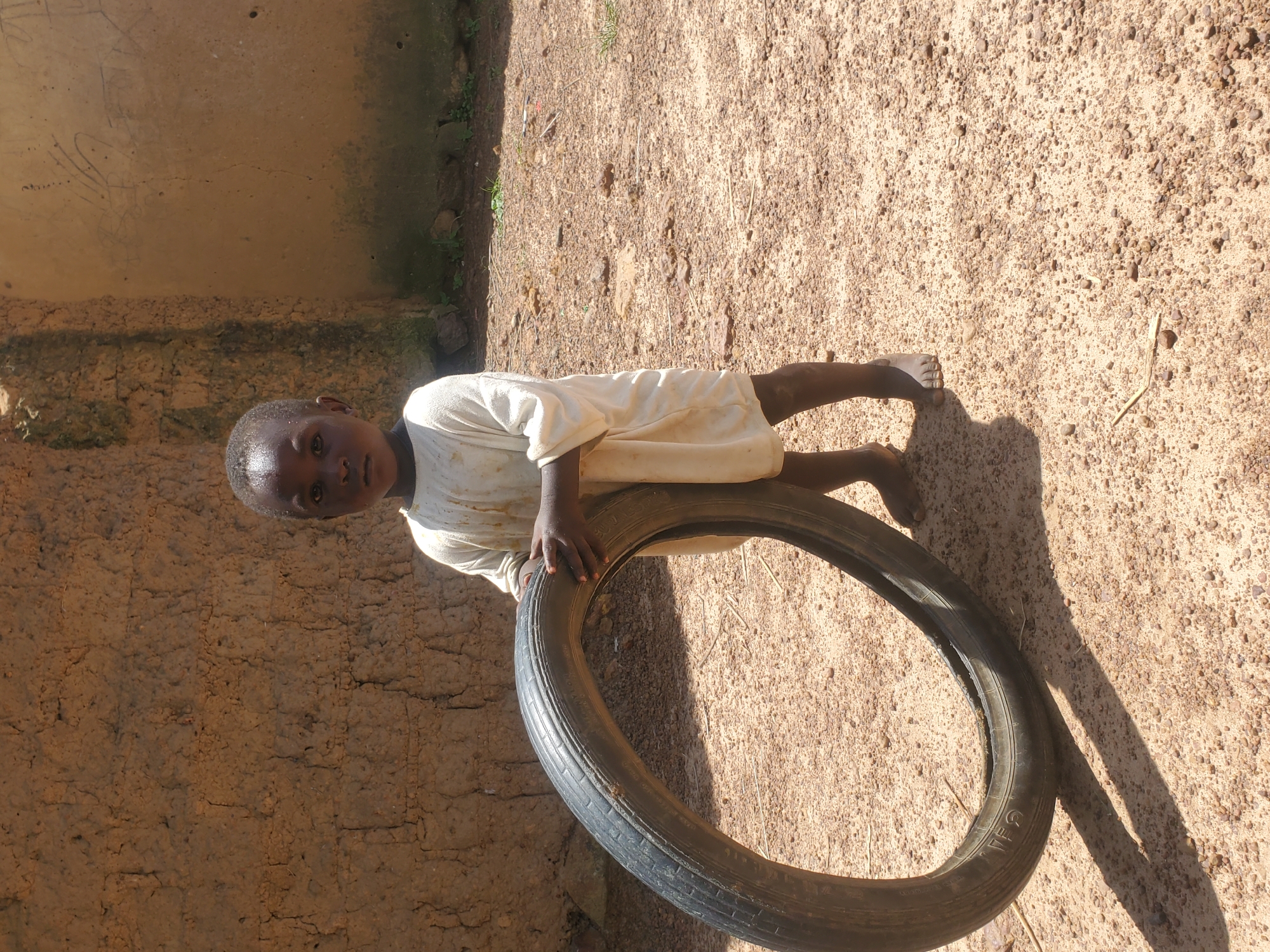 A young child stands outdoors holding a large, black tire. The child is wearing a loose-fitting, light-colored garment and appears to be barefoot. The ground is covered in sand and small stones, with some dry vegetation scattered about. Behind the child, a rough, textured wall, likely made of mud bricks, forms the backdrop. The wall has some rough markings on its surface. The overall scene suggests an outdoor environment in a rural or undeveloped area. The image is taken in Wumat Central, Nigeria.