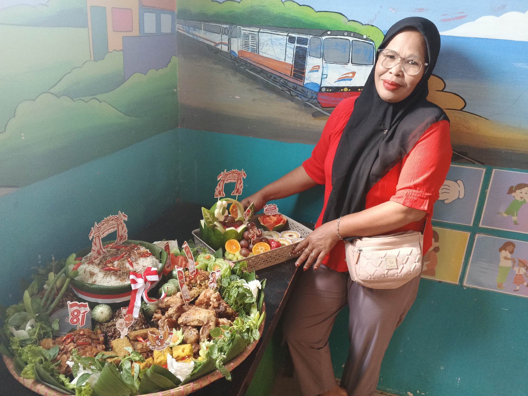 In Warungkiara, Indonesia, a woman wearing a red shirt and a black hijab stands in front of a wall mural depicting a train. She is positioned to the right of two large platters of food. The platter in the foreground is round and filled with rice, fried chicken, vegetables, and various side dishes. It is decorated with small flags and ribbons. Behind it, another platter holds a variety of fruits and what appears to be a carved watermelon. The woman is holding a small decorative item above the fruit platter. The environment suggests an indoor setting, possibly a school or community center, given the presence of the mural and the food display.