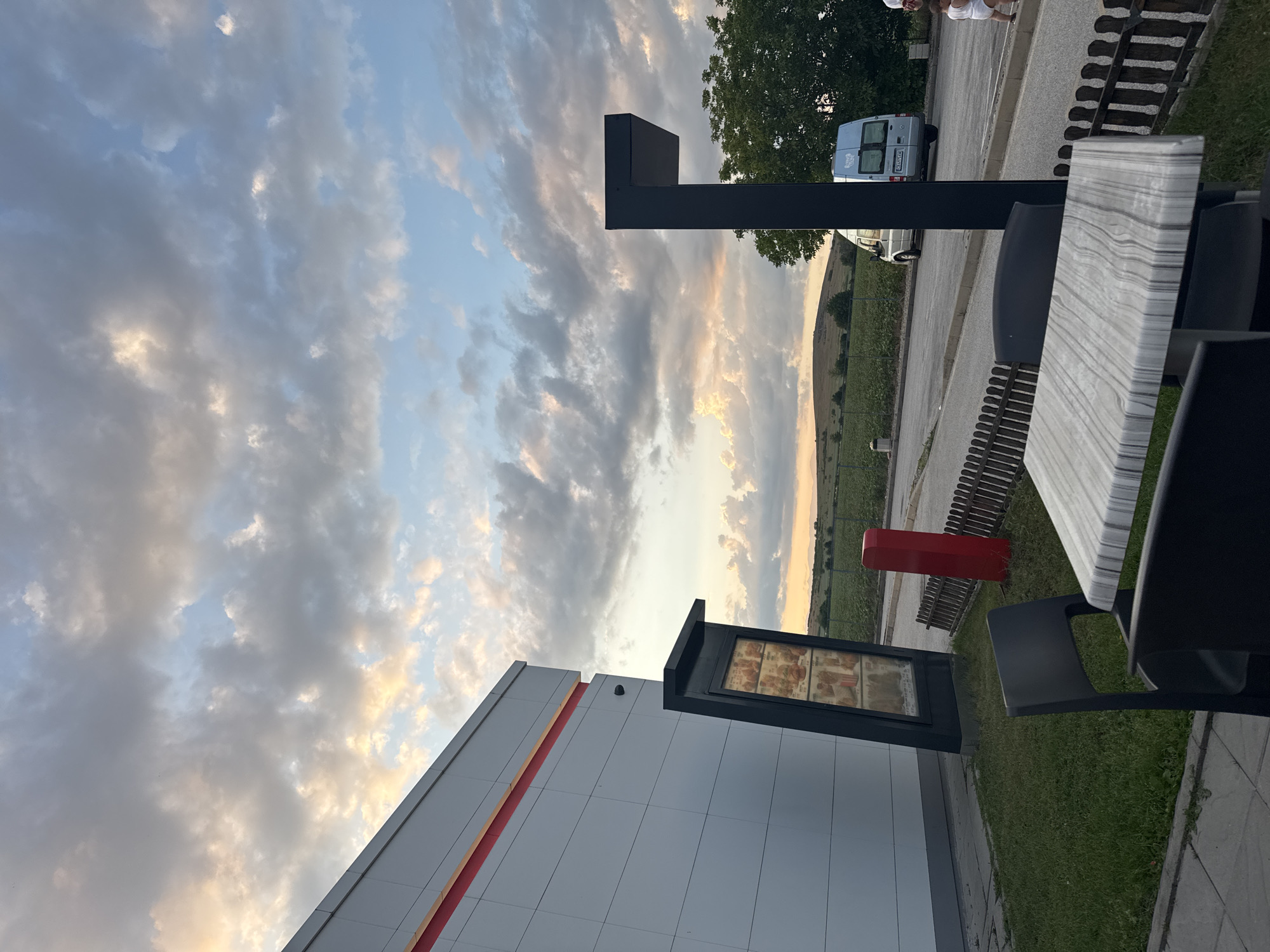 The image captures an outdoor dining area of a commercial establishment in Belozem, Bulgaria, under a cloudy sky at dusk. The sky features a mix of grey and white clouds, with warm golden light from the setting sun concentrated on the left, fading into cooler blue tones on the right.
To the left, a modern building with light grey horizontal panels and a prominent red stripe near the top corner is visible. A dark security camera is mounted on the upper left side of this building. In front of the building, a tilted, black, illuminated menu board displays various food items.
In the foreground, a rectangular outdoor table with a grey and white marble-patterned surface is positioned, accompanied by at least two dark-colored chairs. The immediate ground is paved, transitioning to a strip of green grass. A low, dark wooden picket fence delineates the boundary between the seating area and a paved road beyond. Behind the fence, a tall, slender red post stands to the left.
An asphalt road spans the mid-ground. On the right side of the road, a light-colored Ford Transit van with a partially visible Bulgarian license plate (including 'CA') and a blue sticker on its rear door is either parked or moving slowly, facing away from the viewer. A tall, black, L-shaped pillar or structure is prominently located to the right of the table, partially obscuring the background.
The background reveals extensive green fields of crops and gently rolling hills stretching towards the horizon. A cluster of dark green trees is visible behind the black pillar and to the right of the van. On the far right, a person dressed in a white shirt and shorts is partially visible, accompanying a smaller individual, likely a child or toddler wearing a white diaper or shorts, who is walking on the pavement next to the road.