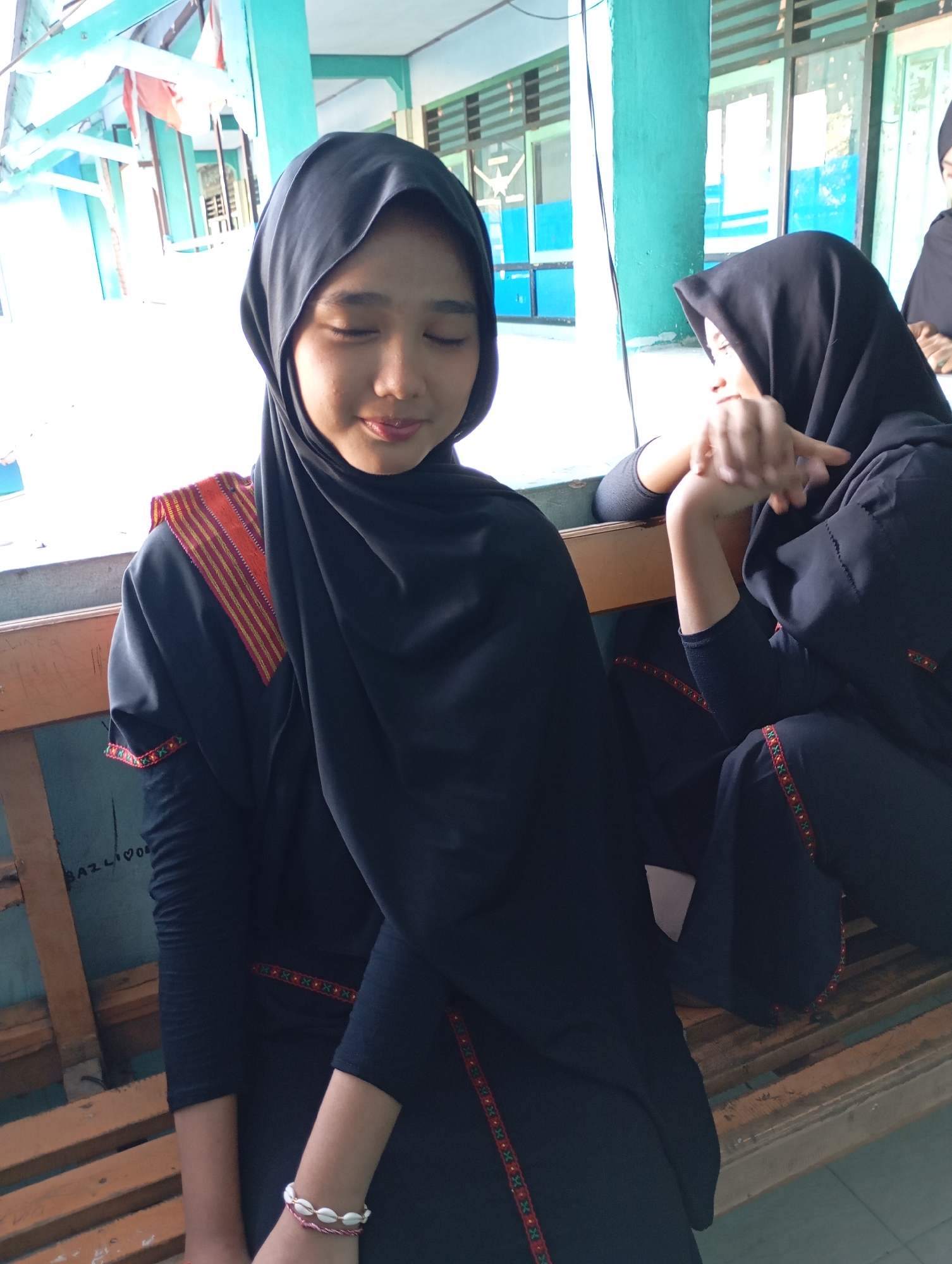 Two young women, likely students, are seated on a wooden bench. The woman in the foreground, wearing a black hijab and black clothing with red and gold trim, has her eyes closed and a slight smile. She is wearing a bracelet made of shells. The woman in the background, also wearing a black hijab and black clothing, is turned away from the camera, with her hands clasped. They appear to be in an outdoor setting with a building structure behind them, possibly part of a school or community center. The lighting suggests it is daytime. The context of Mataram, Indonesia, implies a cultural setting where this attire is common.