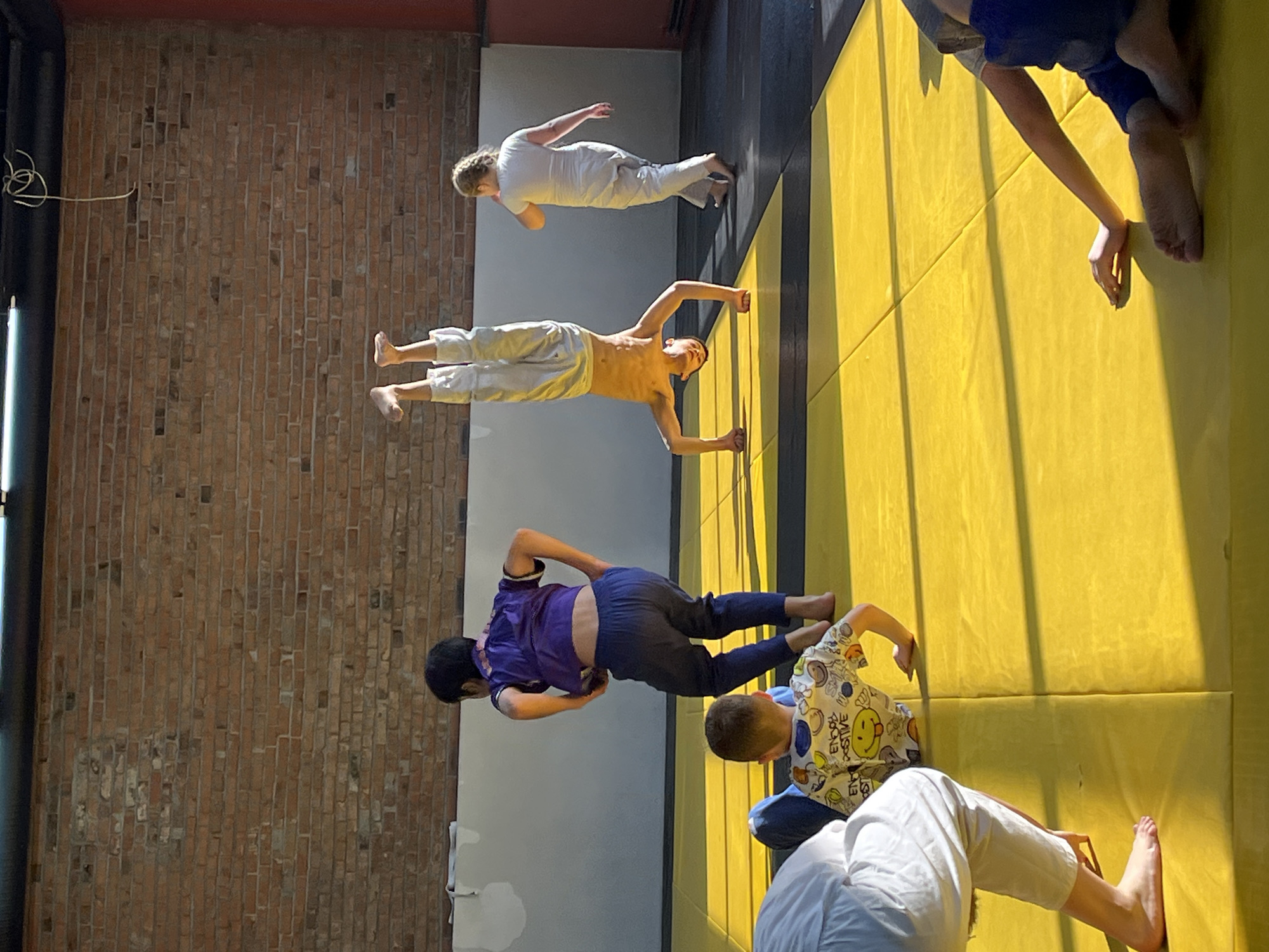 A photograph shows five people lying on a yellow and black padded floor. From left to right, the first person is wearing a purple top and dark pants. The second person is shirtless and wearing light-colored pants. The third person is wearing white clothing. The fourth person is wearing a white top with a pattern and blue pants. The fifth person is wearing white clothing. A brick wall is on the left side of the image. The scene takes place in a gym or martial arts studio. The location might be Krasnoyarsk, Russia.