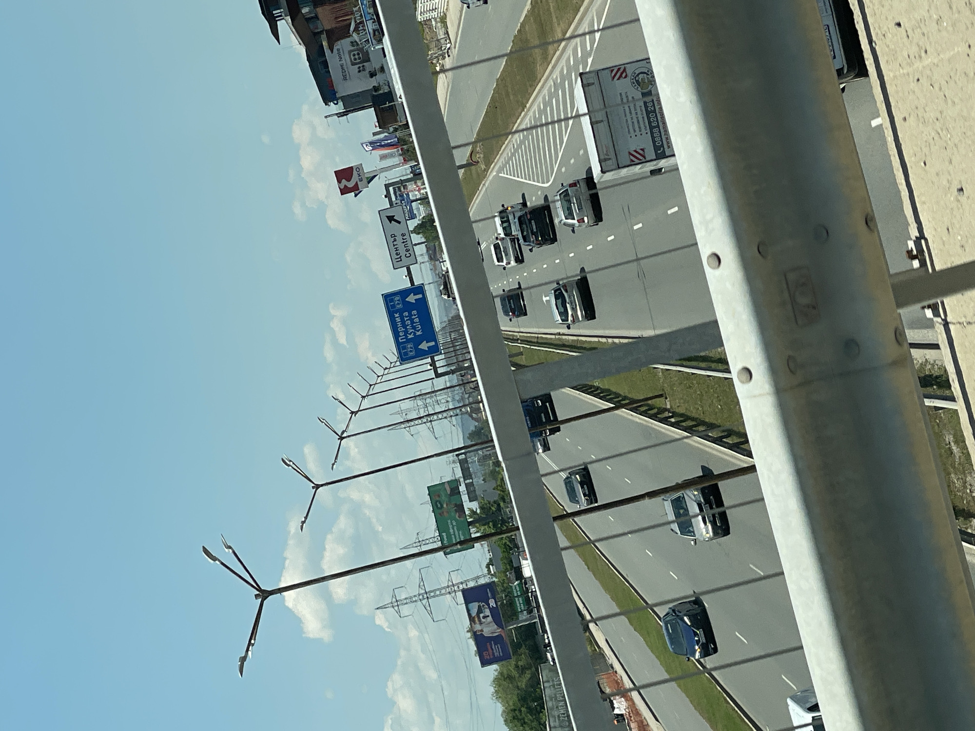 The image depicts a highway scene in Sofia, Bulgaria, on a sunny day with a clear blue sky and scattered clouds. In the foreground, a metal guardrail runs across the frame, obscuring the lower portion of the highway. Beyond the guardrail, a multi-lane highway is visible, with several cars and a truck driving on it. The road is well-maintained, with clear lane markings. On either side of the highway, there are green grassy verges and some trees. In the background, there are buildings, billboards, and power lines, suggesting an urban or suburban environment. One billboard advertises "Zemen Pellets," and another has a "REDMI Note 10" logo. There are also road signs indicating directions to "Pernik," "Kulata," and "Center." Several lampposts are positioned along the highway. The overall impression is of a typical highway commute in a Bulgarian city.