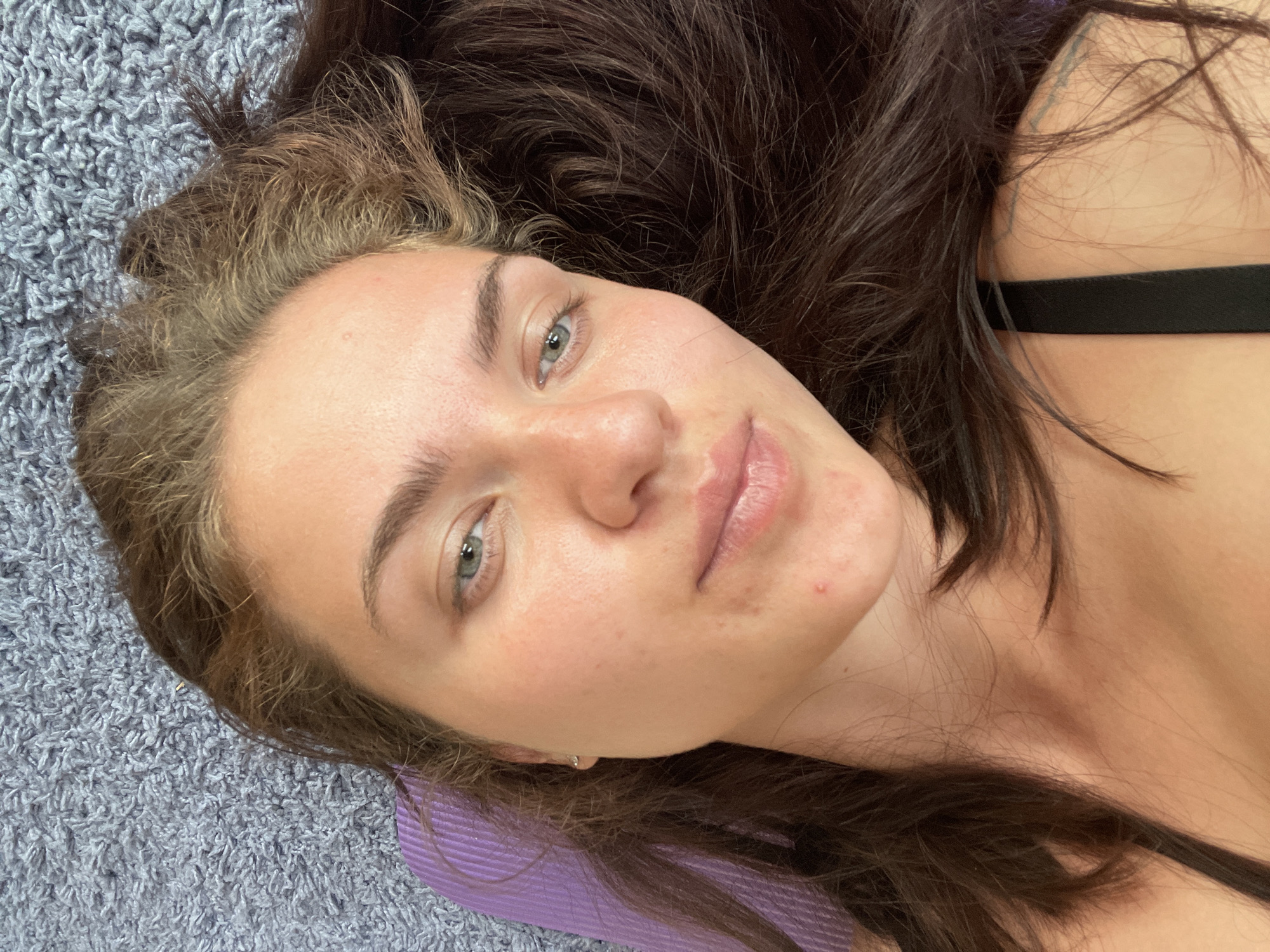 A woman with light skin, light eyes, and a mix of dark brown and lighter brown hair is lying down. She is positioned on a surface that appears to be a combination of a gray shaggy rug and a purple ribbed mat, with her head resting on the mat. Part of a black strap is visible near her shoulder. Her hair is spread out around her head. Her face is visible and is directed towards the camera, and her expression is relaxed. No specific information indicates that the image was taken in the city of Мосметаллоконструкция Онт, Russia.