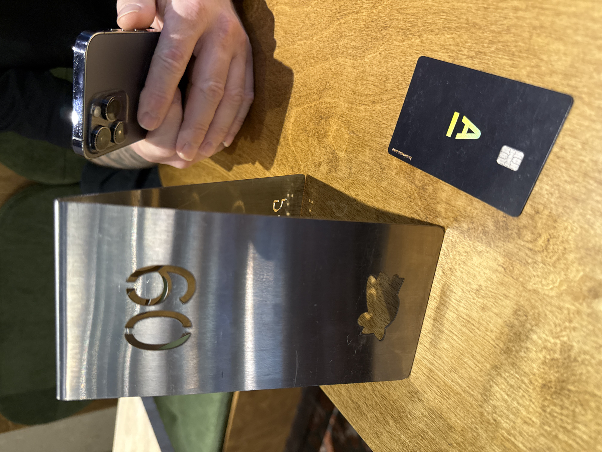 A high-angle shot captures a table scene in what appears to be a restaurant or cafe. A person holds a newer model iPhone, possibly an iPhone 13 Pro or later, in their hands. Next to their hands is a black credit card with a yellow angled "A" logo and the text "business one" visible in the lower left. A metal table number holder displaying "60" and a small, stylized bear shape cut out is present. The table is wooden with a visible grain. The scene lacks specific identifying details to confirm its exact location in Sochi, Russia, but the general environment suggests a modern dining establishment.