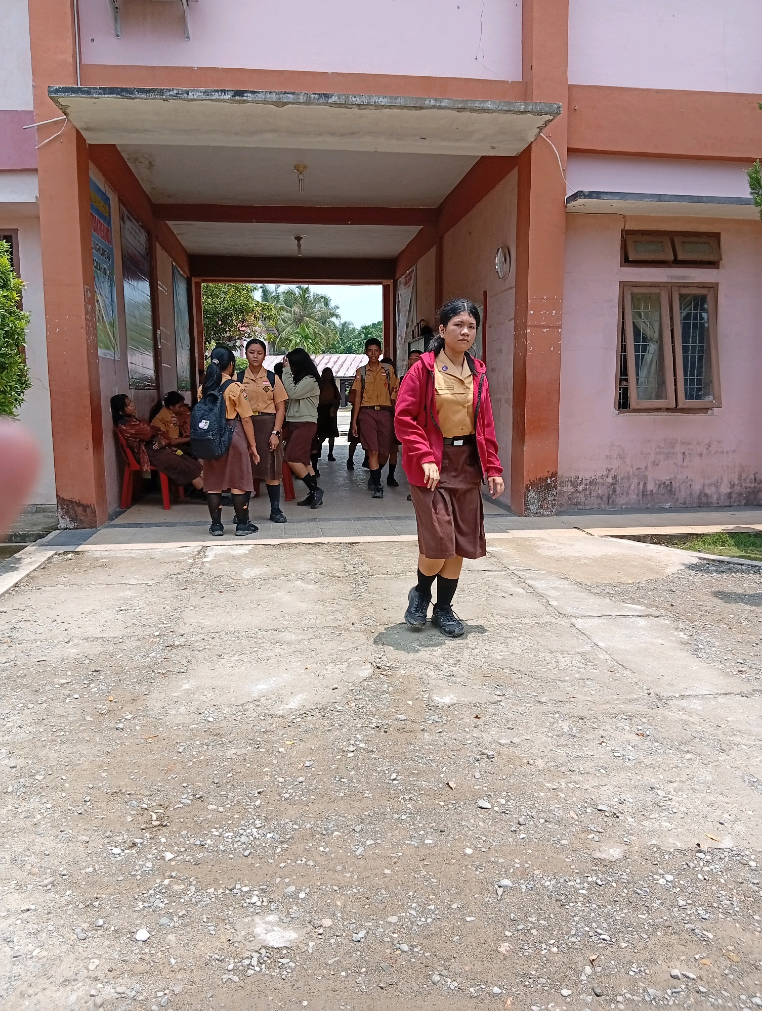 In the city of Gunungsitoli, Indonesia, a group of young people in school uniforms are seen standing and walking in front of a building. The building has a pink facade and a covered walkway with an orange trim. Several students are gathered in the walkway, some sitting on chairs and others standing. In the foreground, a young woman wearing a red jacket over a tan school uniform and a brown skirt walks forward, looking towards the camera. The ground is paved with a mix of concrete and gravel. The overall scene appears to be a school campus or a similar educational institution.