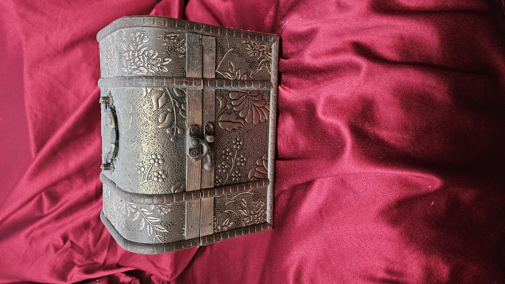 A decorative wooden chest with embossed floral and grape vine patterns sits on a crumpled red velvet fabric. The chest is rectangular with rounded edges and a hinged lid, secured by a metal clasp. The fabric underneath displays folds and highlights characteristic of velvet, with variations in light and shadow. The scene is static and appears to be a still life composition.