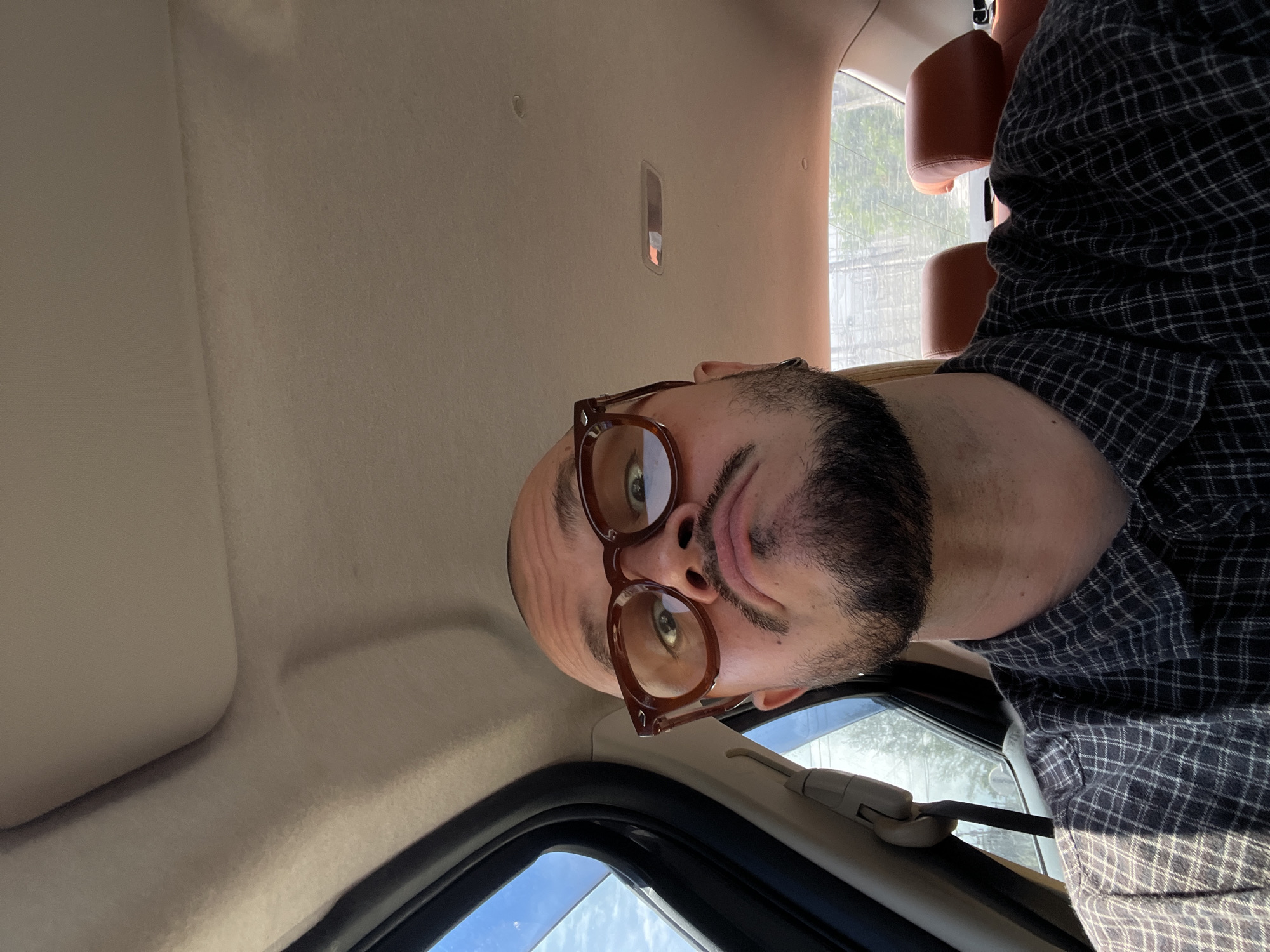 A close-up, head-and-shoulders selfie of a bald, white man with a beard and glasses is taken from a low angle inside a vehicle. The man is wearing a dark, checkered shirt and is looking directly at the camera with a puckered expression. The interior of the car, including the headliner and leather seats, is visible. The man's head is slightly tilted to the left, and his glasses have thick, brown frames. The lighting suggests it is daytime, with sunlight entering through the car window.