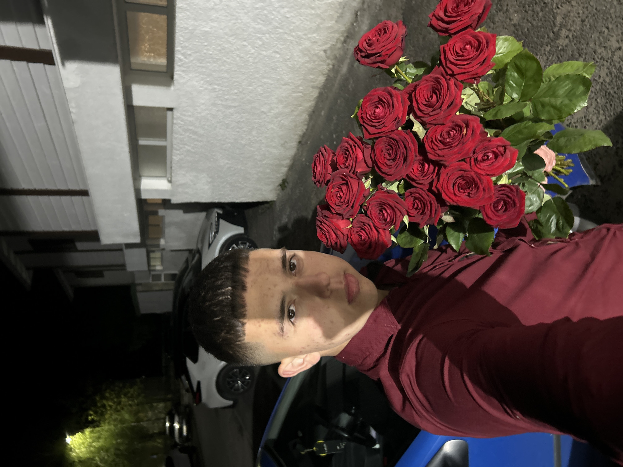 The image shows a young man holding a bouquet of red roses. He is wearing a burgundy-colored shirt and is positioned in front of a blue car. In the background, there is a building with visible windows and parked cars. The location is likely outdoors during the evening. The specific location within Zlatograd, Bulgaria cannot be determined from the image.