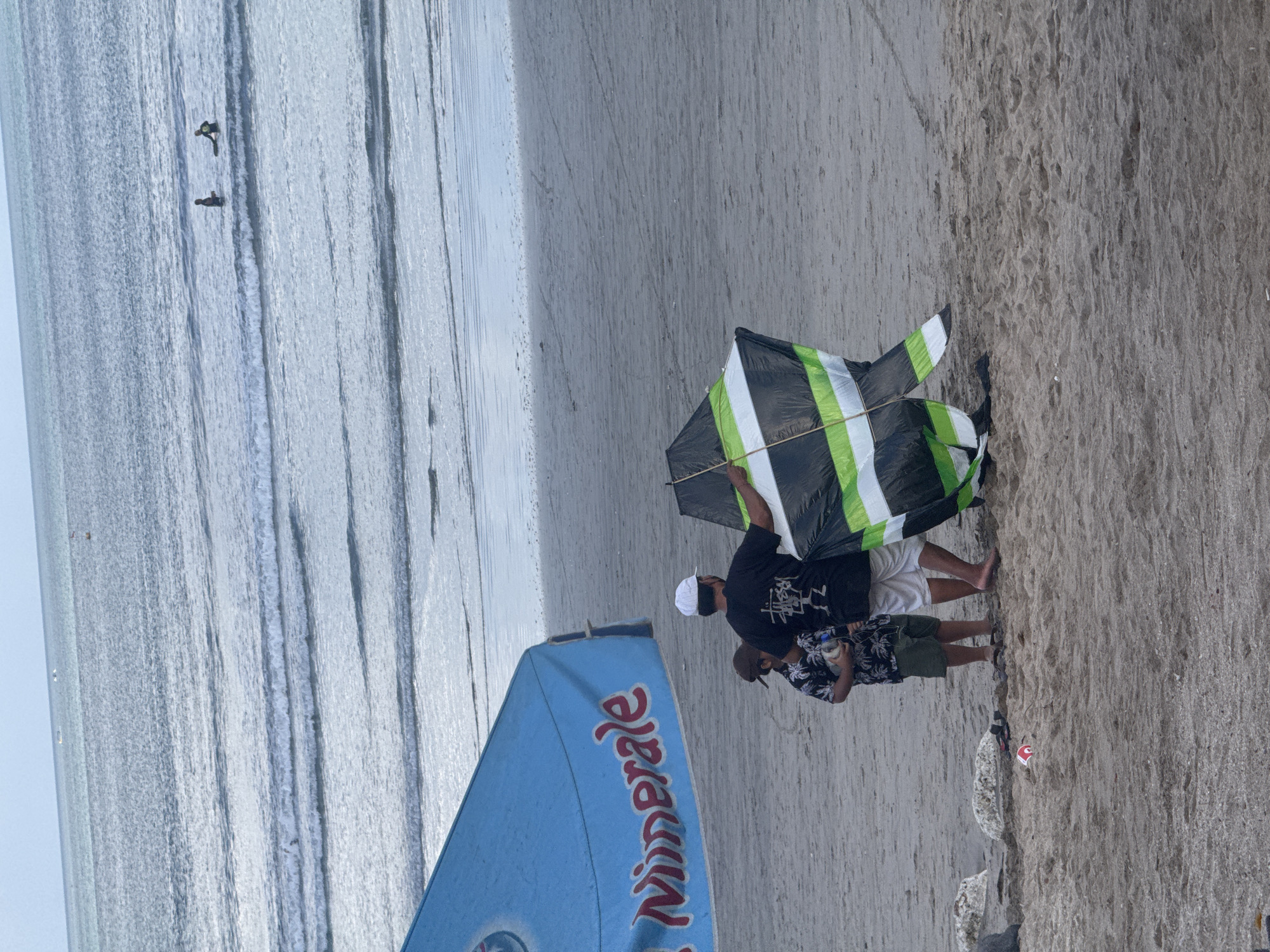 A man wearing a white cap and a black t-shirt stands on a sandy beach in Kuta, Indonesia, holding a black, green, and white striped kite. Next to him, a child in a dark patterned shirt and green shorts stands looking at the kite. In the background, the ocean has small waves, and two people are in the water, one with a surfboard. A large blue tarp with red lettering that reads "Minerale" is visible in the foreground to the left. The ground is covered in sand, with some rocks and a discarded red cup visible.