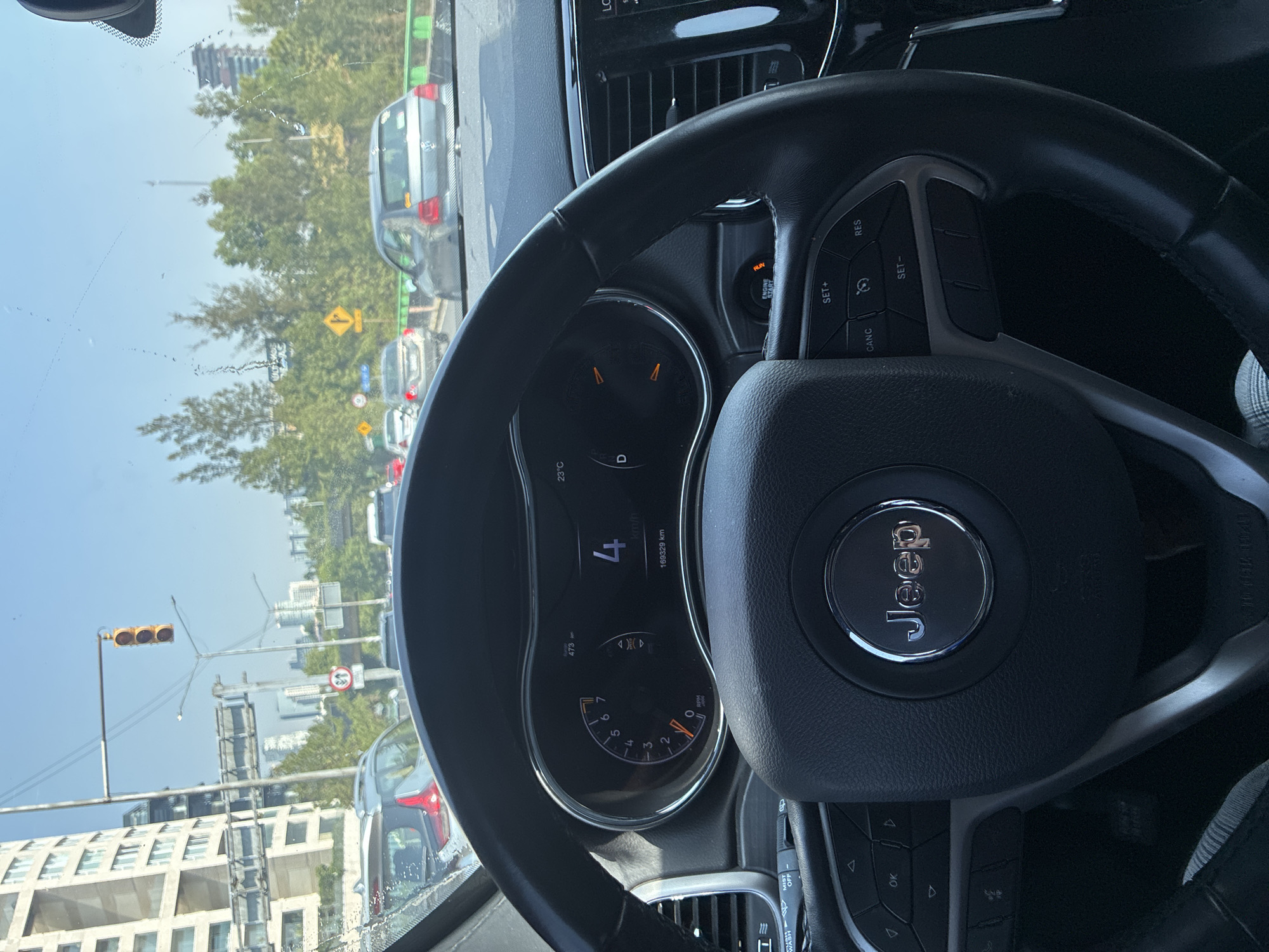 The image shows the interior of a Jeep vehicle, focusing on the steering wheel and dashboard. The speedometer displays a speed of 4 km/h and a total mileage of 169,329 km. The temperature is 23°C. Outside the vehicle, a cityscape is visible, including traffic lights and several buildings, with dense traffic congestion. The location is Mexico City, Mexico.