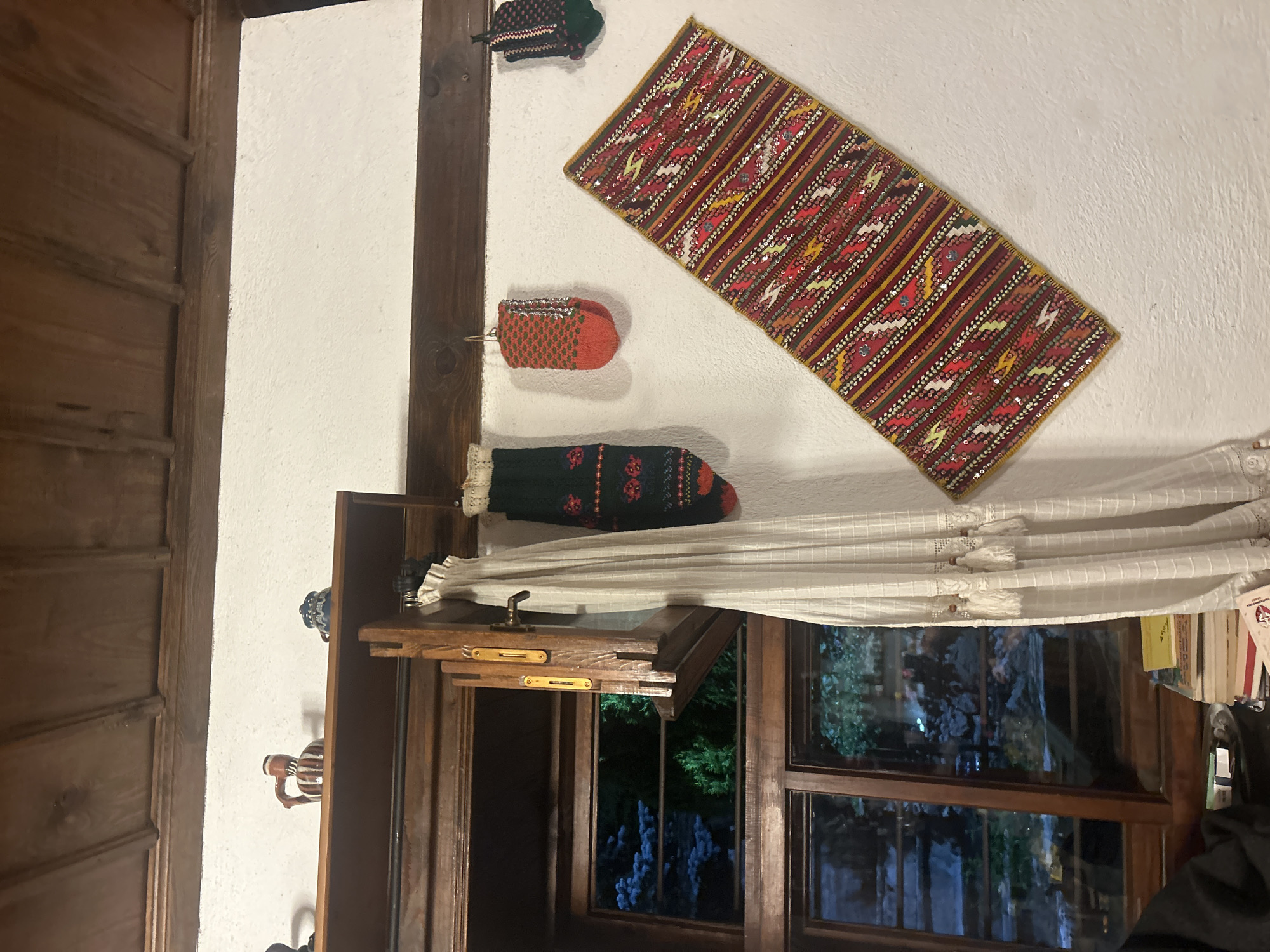 A vertical, medium shot taken indoors in Dolen, Bulgaria, features a white wall adorned with traditional Bulgarian crafts: a woven runner with red, gold, and green patterns; three decorative sock-like items in red, green, and brown; and a white curtain with tassels hanging beneath them. Below, a shelf rests above a window that reveals a dark scene outside. The left side of the frame shows wooden paneling, and the window offers a view of foliage and distant lights.