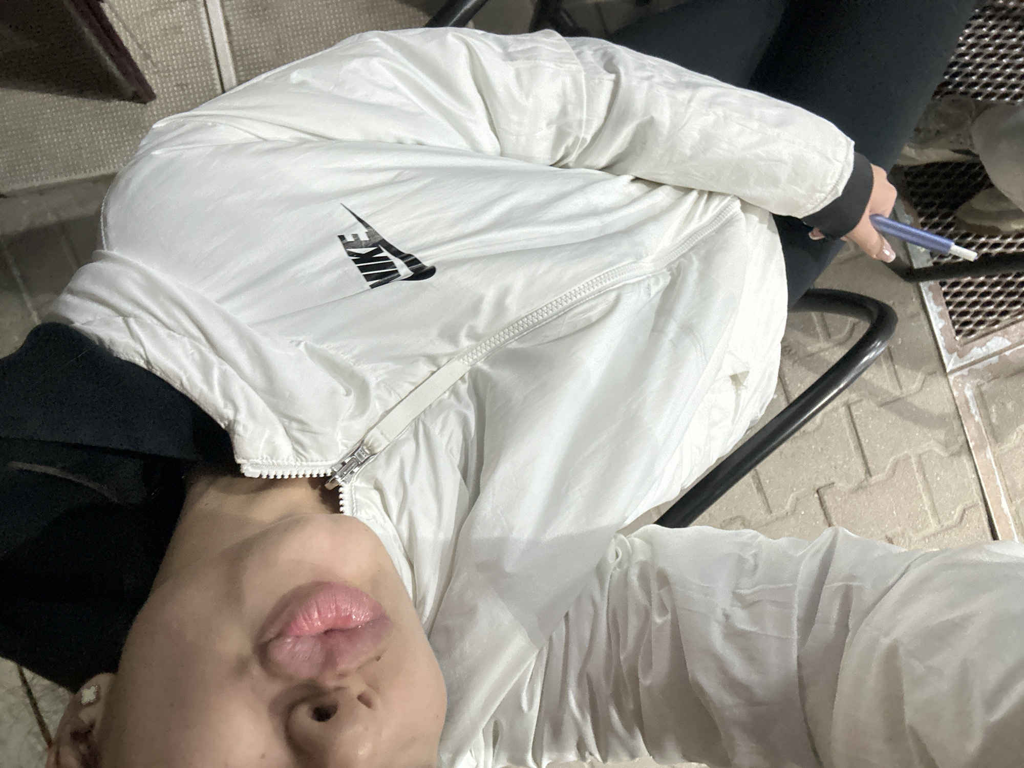 A selfie, taken from a slightly overhead perspective, captures a person wearing a white Nike puffer jacket. The jacket is partially zipped and features the Nike logo prominently on the left breast. The person's face, including their lips, nose, and part of their chin, is visible in the lower portion of the frame. The subject is seated in a black chair, the frame of which is visible around them. They are holding a blue pen-like object in their right hand. The background appears to be an outdoor area, possibly a patio or balcony, with a textured surface resembling tiles or grating. The scene is illuminated with artificial lighting, suggesting it might be nighttime. Without additional cues, it is not possible to confirm the location in Plovdiv, Bulgaria.