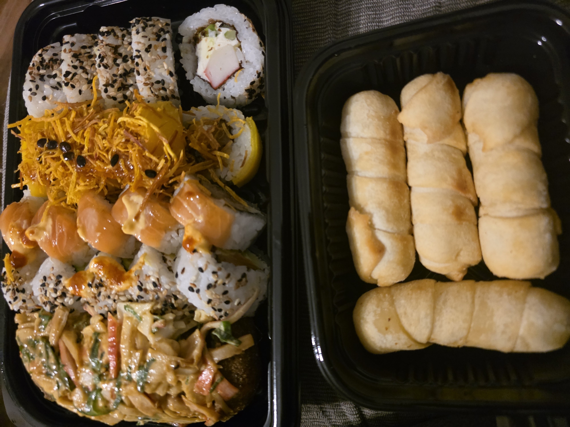 The image displays two black plastic takeout containers of food positioned on a dark, textured surface, likely a tablecloth.
The top container, larger in size, holds an assortment of sushi rolls. From left to right: a large, rounded sushi item covered in a creamy, light-brown sauce with visible green and orange julienned vegetables; multiple rows of sushi rolls topped with orange salmon slices and a creamy orange drizzle; a section of rolls covered extensively with thin, golden-yellow crispy strands (possibly fried sweet potato or onion) and dotted with black seeds; and a row of rolls coated primarily in black and white sesame seeds. In the bottom right corner of the sushi arrangement, a single maki roll is visible, showing a filling of white cream cheese, green, and a red/pink strip (likely imitation crab). A yellow lemon wedge is situated near the center of the sushi assortment.
The bottom container, smaller and positioned below the first, contains five golden-brown, cylindrical, dough-wrapped items, characteristic of tequeños. One is oriented vertically on the left, while the other four are arranged horizontally to its right.
No individuals, actions, or interactions are depicted. The specific location within the city of Buenos Aires, Argentina, cannot be determined from the visual information provided.