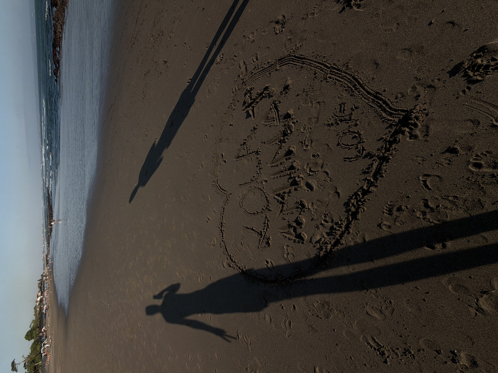 A wide shot captures a dark sand beach in North Kuta, Indonesia, with the ocean in the background under a clear sky. The sun casts long shadows across the sand, with one shadow in the foreground depicting a person taking a selfie and another extending further into the distance. A heart shape is drawn in the sand, containing the words "I Love You" written inside. The beach has scattered footprints and some small waves are visible at the water's edge, with a few distant people and beach umbrellas visible on the shore.
