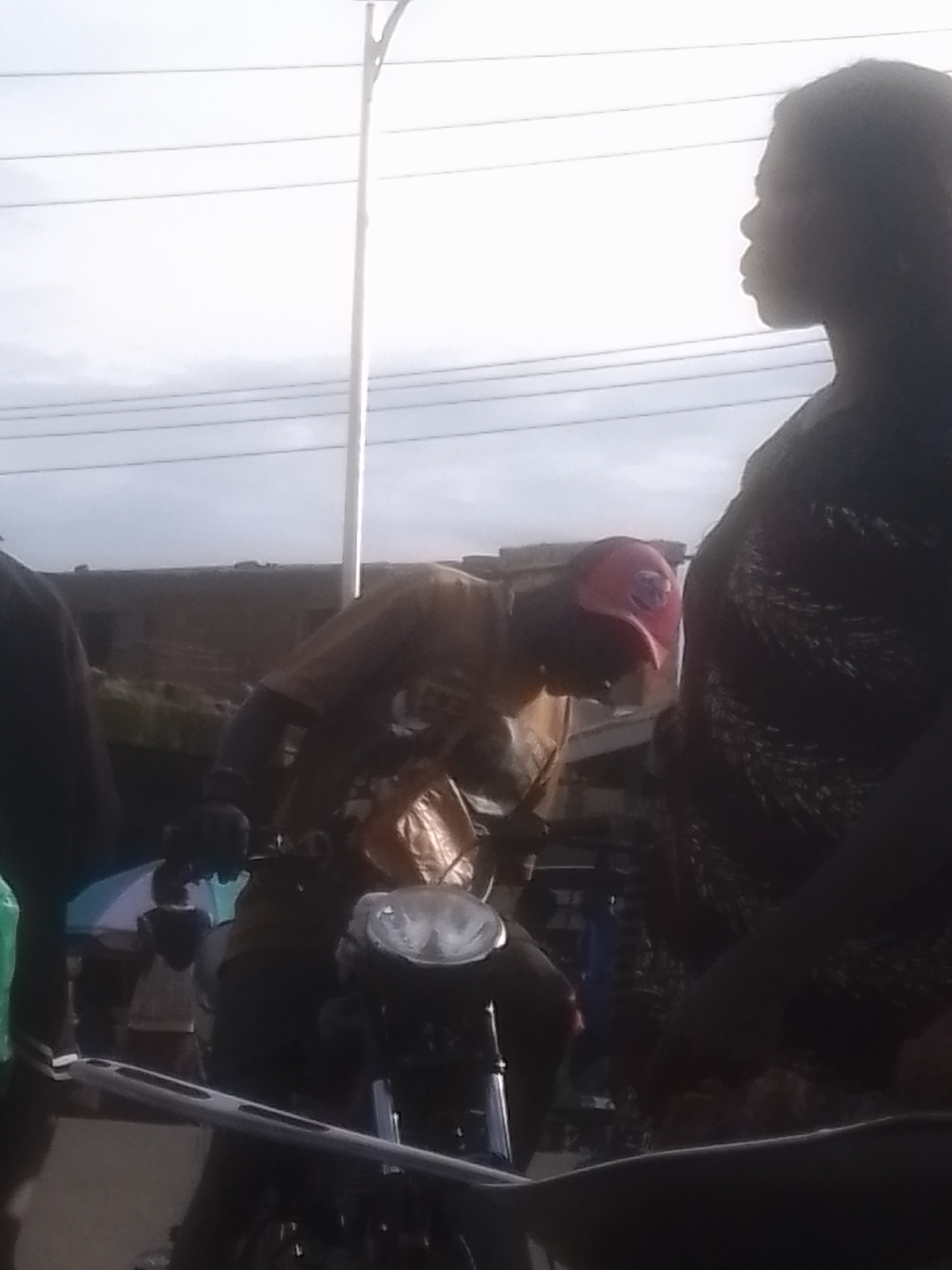 In Gwong, Nigeria, a man wearing a red baseball cap and a tan shirt is seated on a motorcycle, leaning forward. He appears to be interacting with the motorcycle's controls. To the right of the motorcycle, a woman with dark hair is visible in profile, looking to her left. In the background, a low, dark building with a textured facade is present. Power lines are strung across the sky, which is overcast with some clouds. The scene appears to be outdoors, likely on a street or roadside.